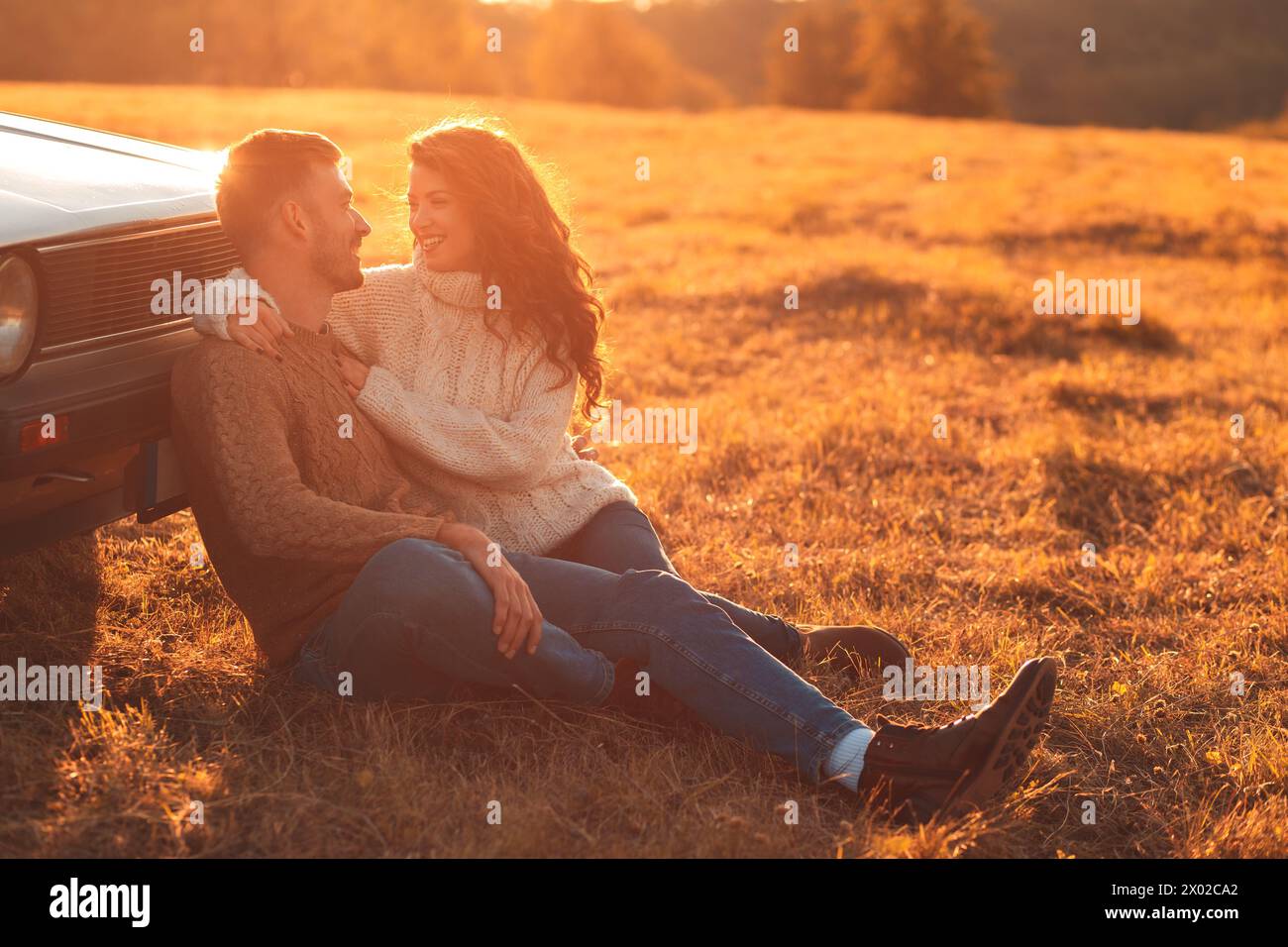 Beau jeune couple profitant du temps ensemble assis en plein air sur la prairie s'appuyant sur la vieille voiture embrassant les uns les autres. Banque D'Images