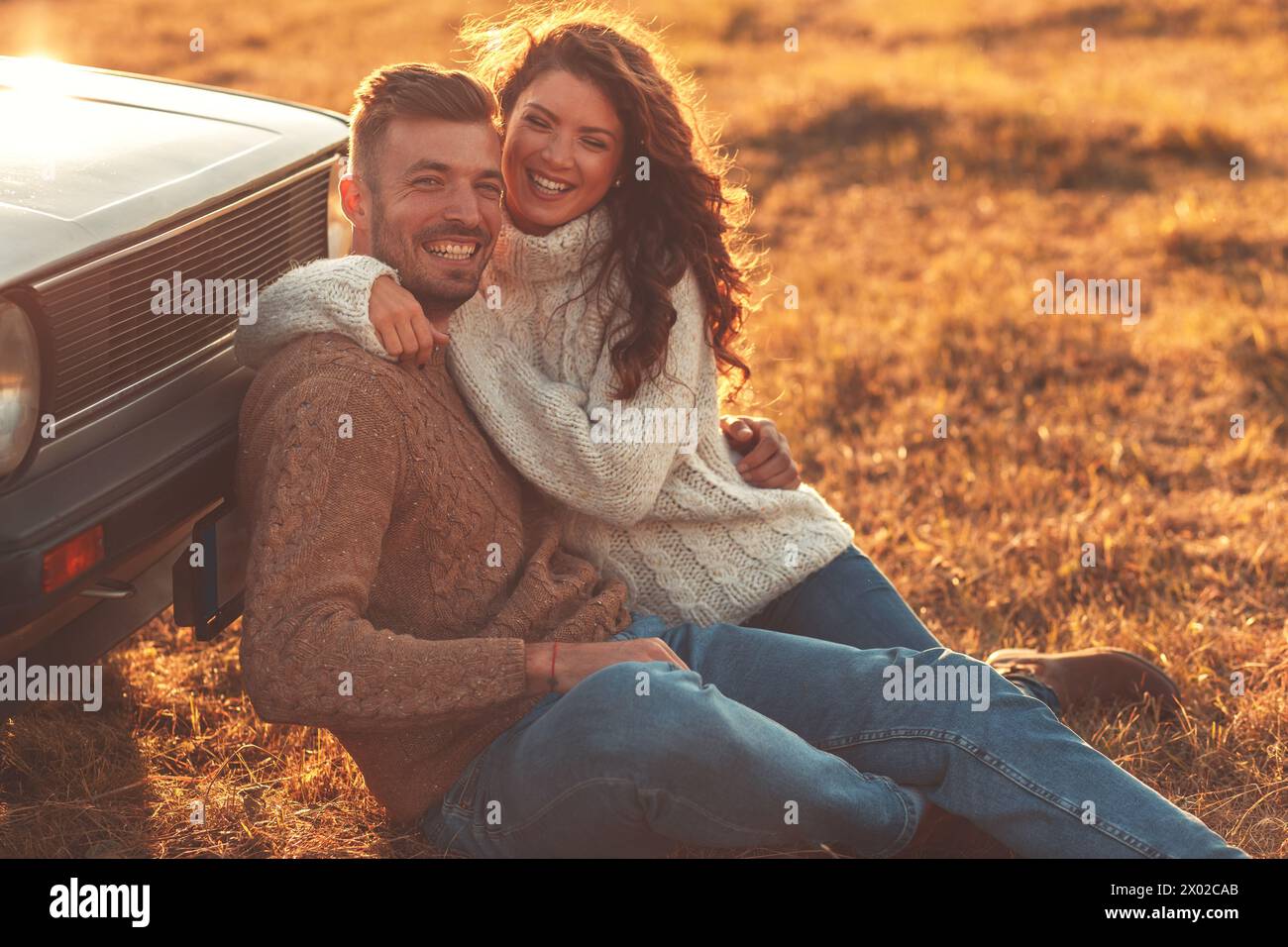 Beau jeune couple profitant du temps ensemble assis en plein air sur la prairie s'appuyant sur la vieille voiture embrassant les uns les autres. Banque D'Images