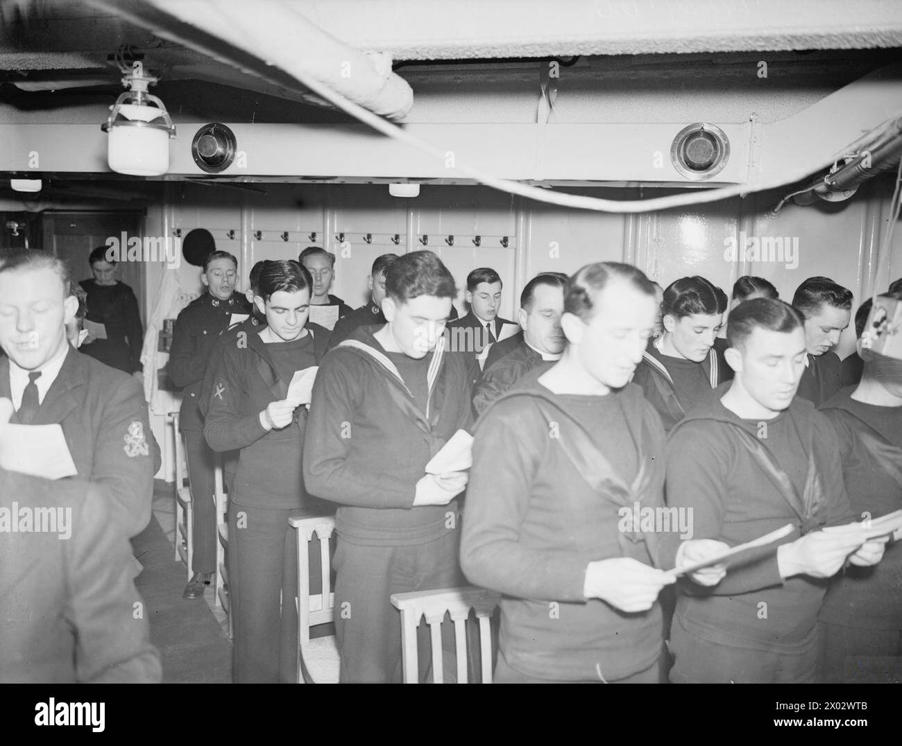 SCÈNES DE NOËL À BORD DU HMS VICTORIOUS. 20 DÉCEMBRE 1941, À BORD DU PORTE-AVIONS DE SCAPA. - Notes participant à un service volontaire de chant de Noël dans l'église à bord du HMS VICTORIOUS qui a été transmis dans tout le navire. Le Padre, Rev Dixon, RN, qui était dans l'AJAX dans la bataille DE GRAF SPEE, peut être vu avec les hommes dans la 2ème rangée, 2ème à droite. Le microphone peut être vu près de la bande de cordes jouée par les Royal Marines Banque D'Images