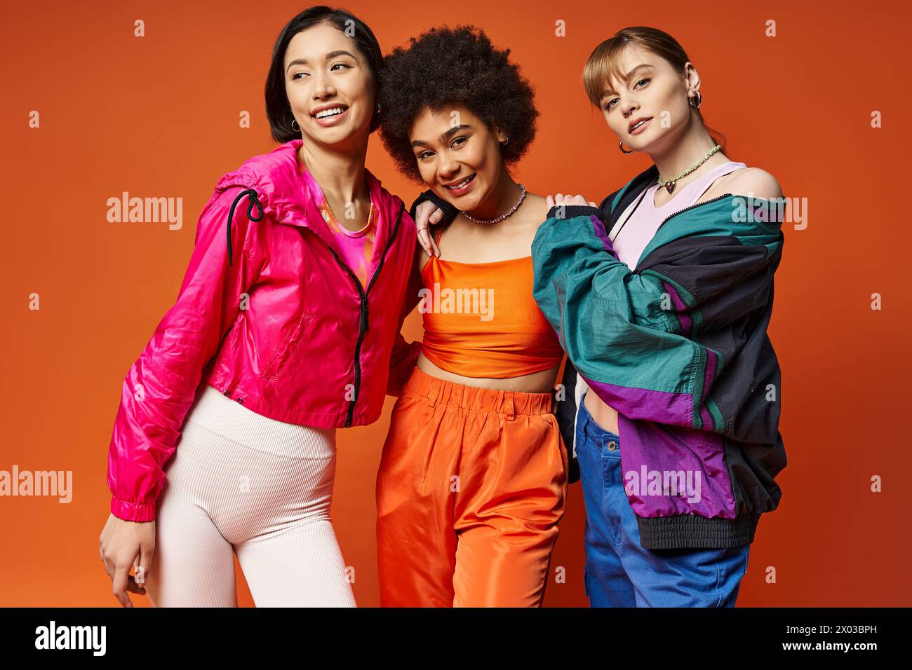 Trois femmes de différentes ethnies se tiennent confiantes dans un studio avec un fond orange, mettant en valeur la beauté multiculturelle. Banque D'Images