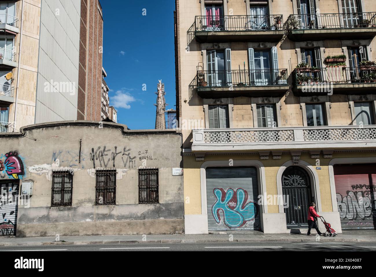 Lady en rouge pousse une poussette dans une rue de Barcelone, avec la Sagrada Familia en arrière-plan parmi les graffitis et les vieux bâtiments. Banque D'Images