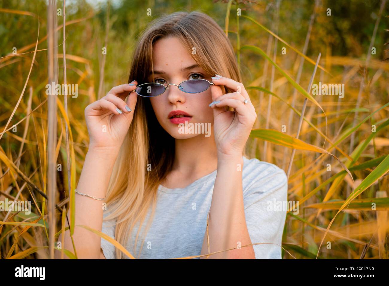 Belle jeune fille portant un t-shirt gris vierge et un Jean noir dans des lunettes bleues posant contre l'herbe haute vert et jaune au début de l'automne chaud. Banque D'Images