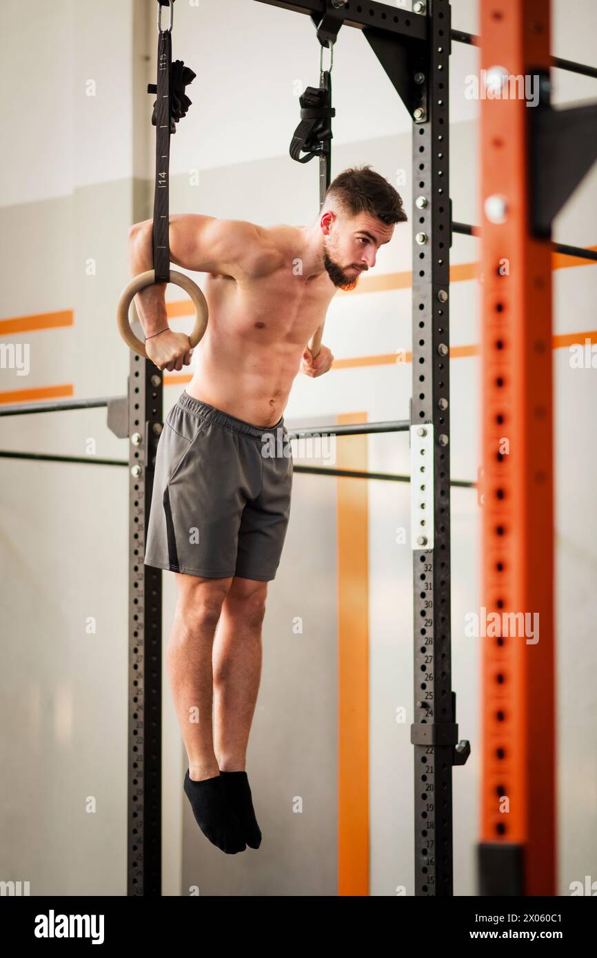 Corps complet de sportif fort en short suspendu et balançant sur des anneaux de gymnastique au gymnase pendant l'entraînement fonctionnel Banque D'Images