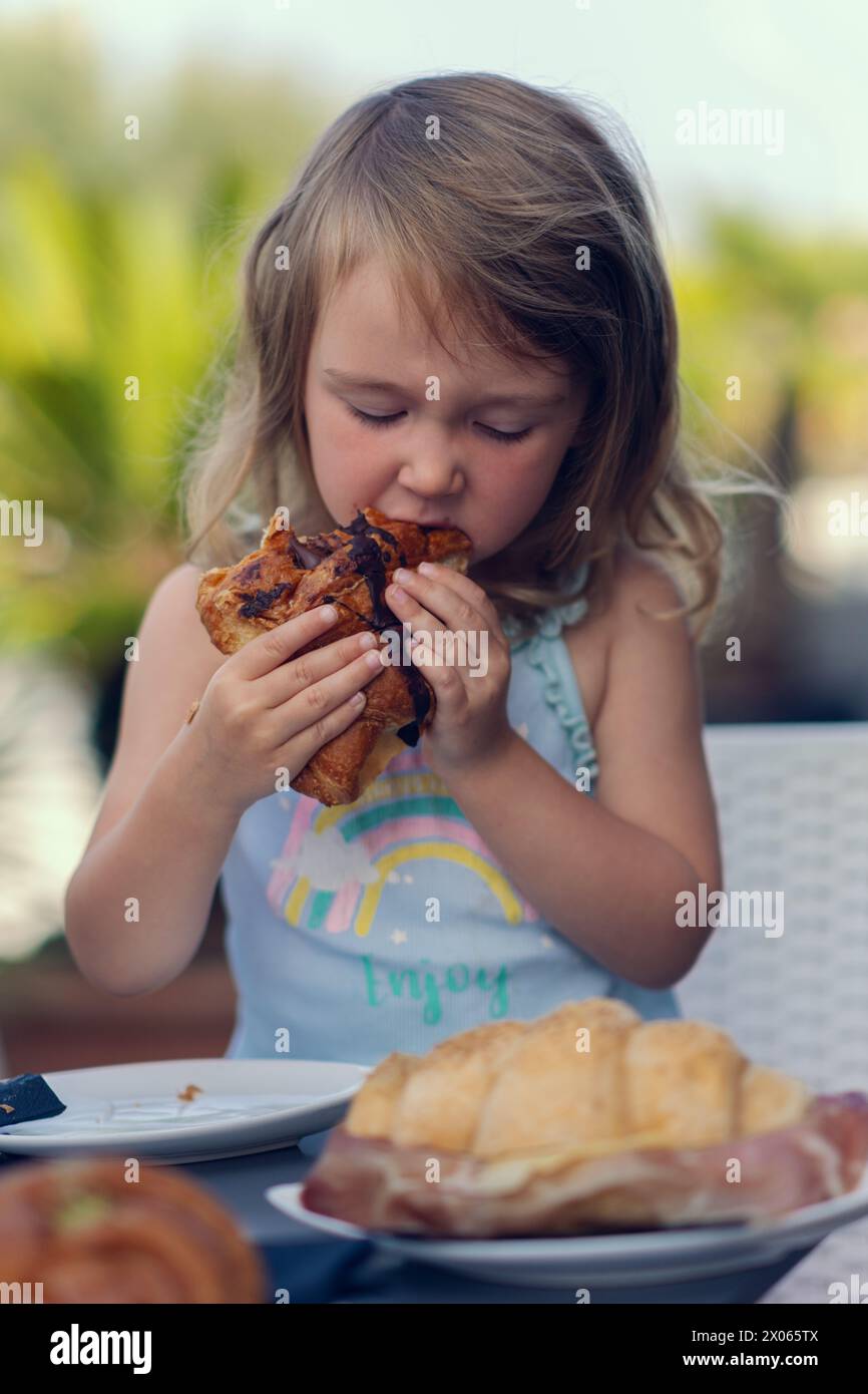 Une belle petite fille mange un croissant avec du chocolat avec de l'appétit. La fille prend son petit déjeuner avec un croissant. Une fille caucasienne mange des pâtisseries. Gros plan Banque D'Images