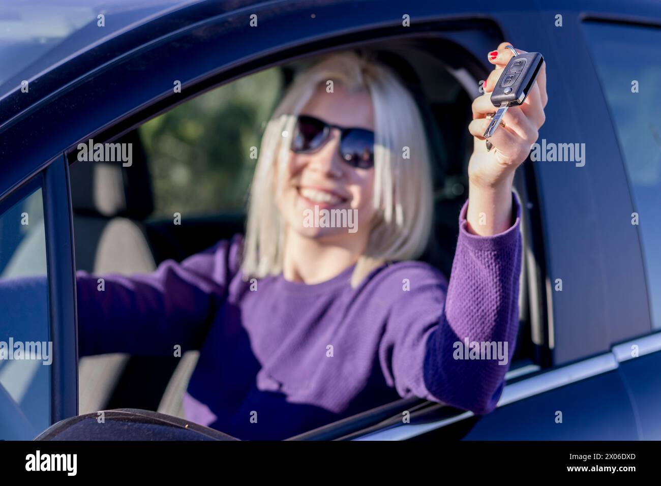 Femme blonde joyeuse dans un pull violet, avec des lunettes de soleil sur sa tête, affichant joyeusement ses clés de voiture, symbolisant une nouvelle propriété ou liberté Banque D'Images