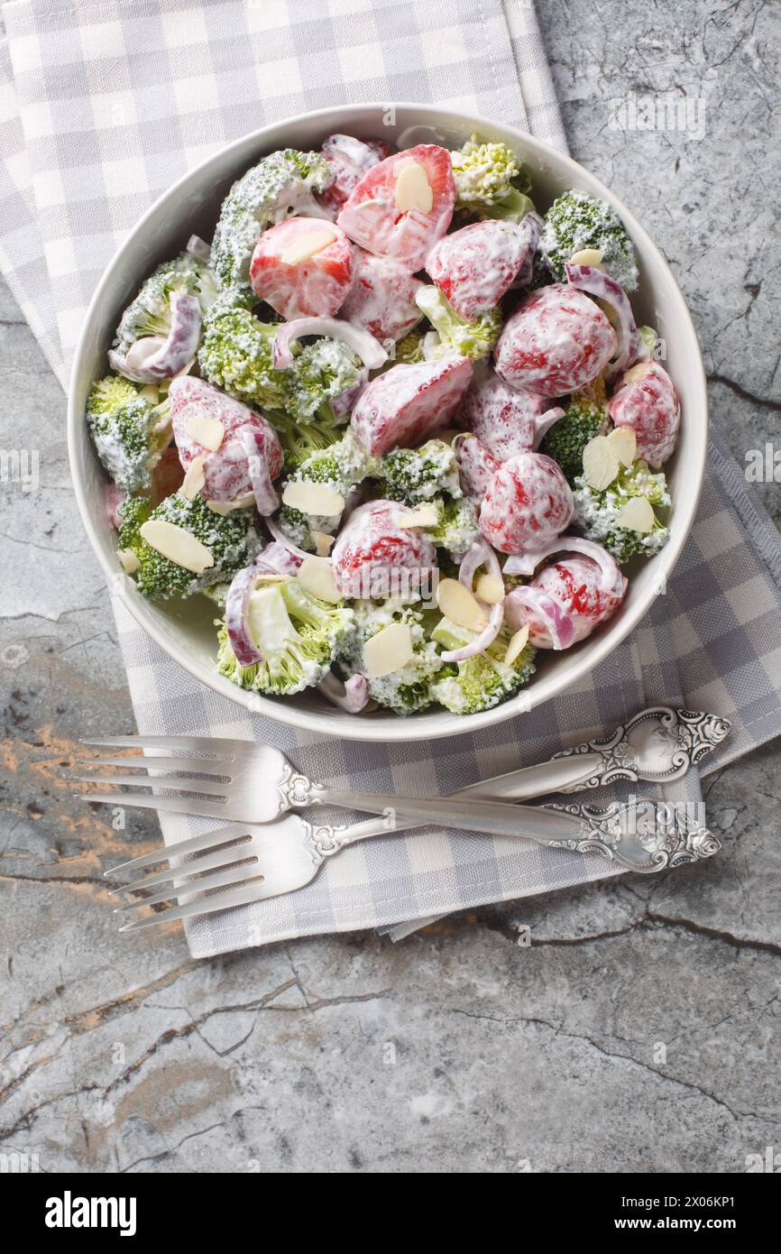 Salade de brocolis frais avec fraises, oignons rouges et amandes habillées de yaourt gros plan dans une assiette sur la table. Vue de dessus verticale Banque D'Images