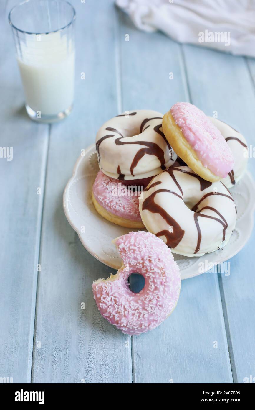Beignets à la vanille givrés avec des tourbillons de chocolat et des beignets roses à la fraise avec des flocons de noix de coco. Morsure manquante. Verre de lait en arrière-plan. Banque D'Images