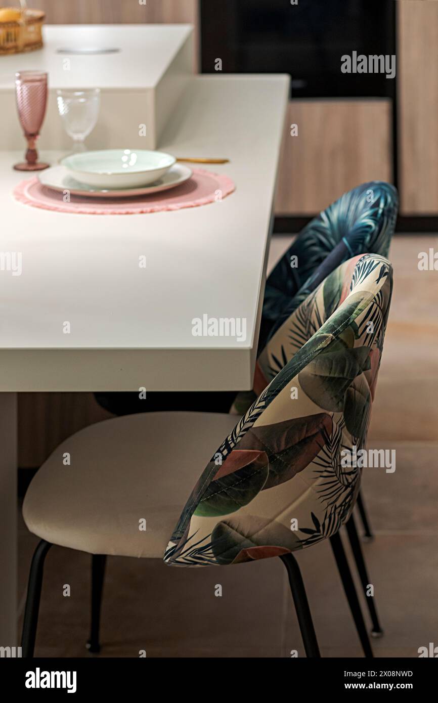 Un coin salle à manger moderne avec un plateau blanc avec un napperon rose, un ensemble de plats, un verre délicat et une chaise unique avec un imprimé tropical Banque D'Images