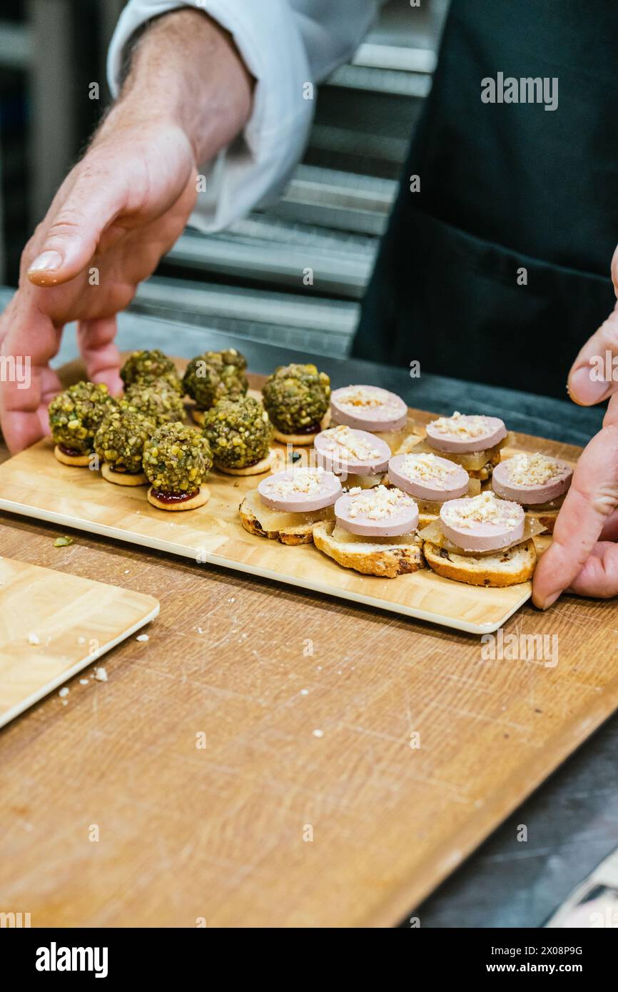 Anonymous Chef Hands arrangeant soigneusement un assortiment de canapés complexes surmontés de pâté et de pistaches sur une planche de service en bois Banque D'Images