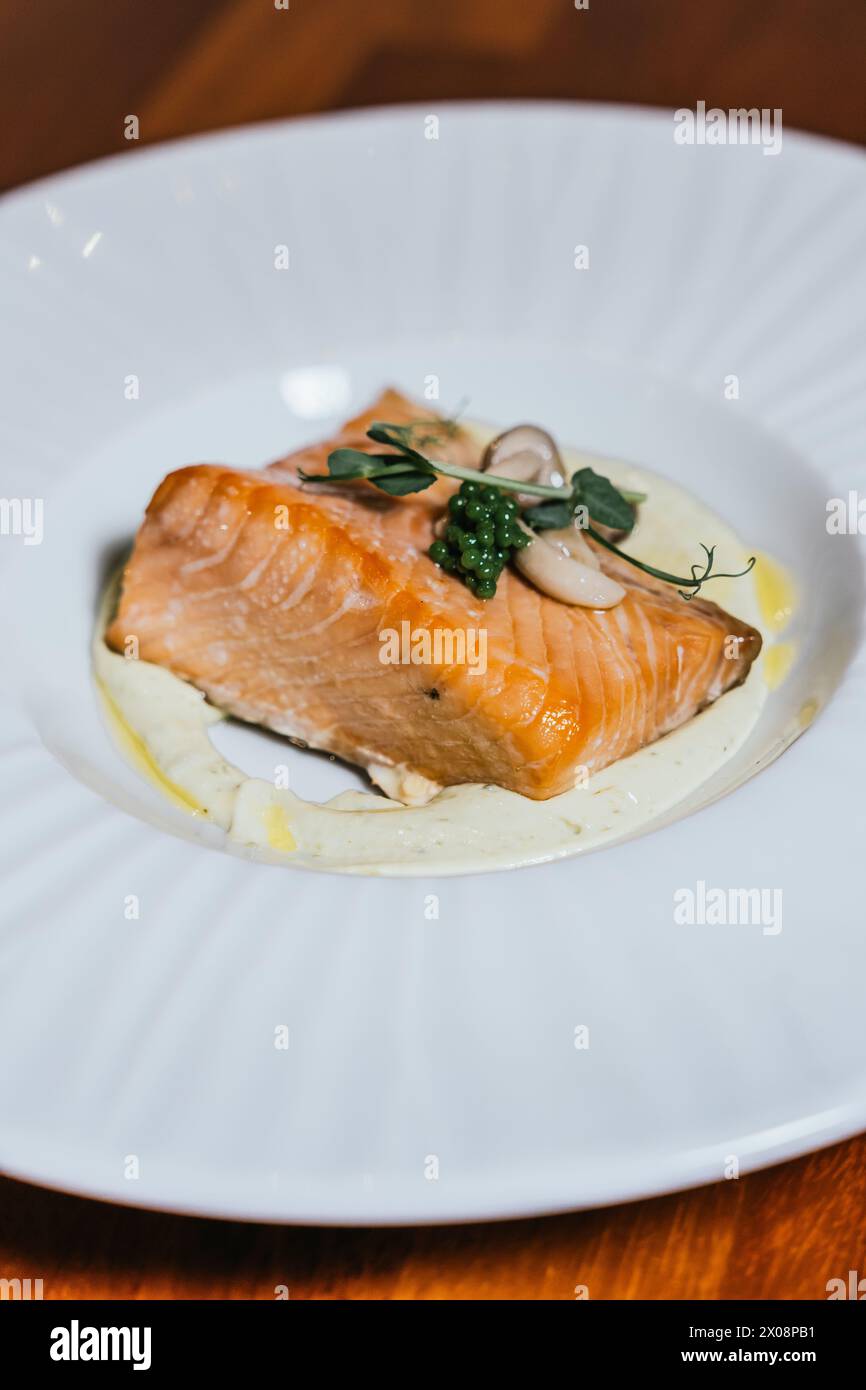 Un filet de saumon magnifiquement poché repose sur une base crémeuse, élégamment garni de caviar et d’herbes fraîches sur une assiette blanche Banque D'Images