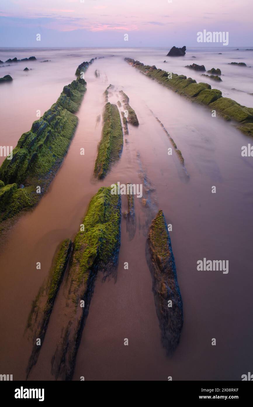 La lumière de l'aube adoucit la scène à Playa de Barrika, Bilbao, où des formations rocheuses couvertes de mousse créent un sentier naturel vers la mer Banque D'Images