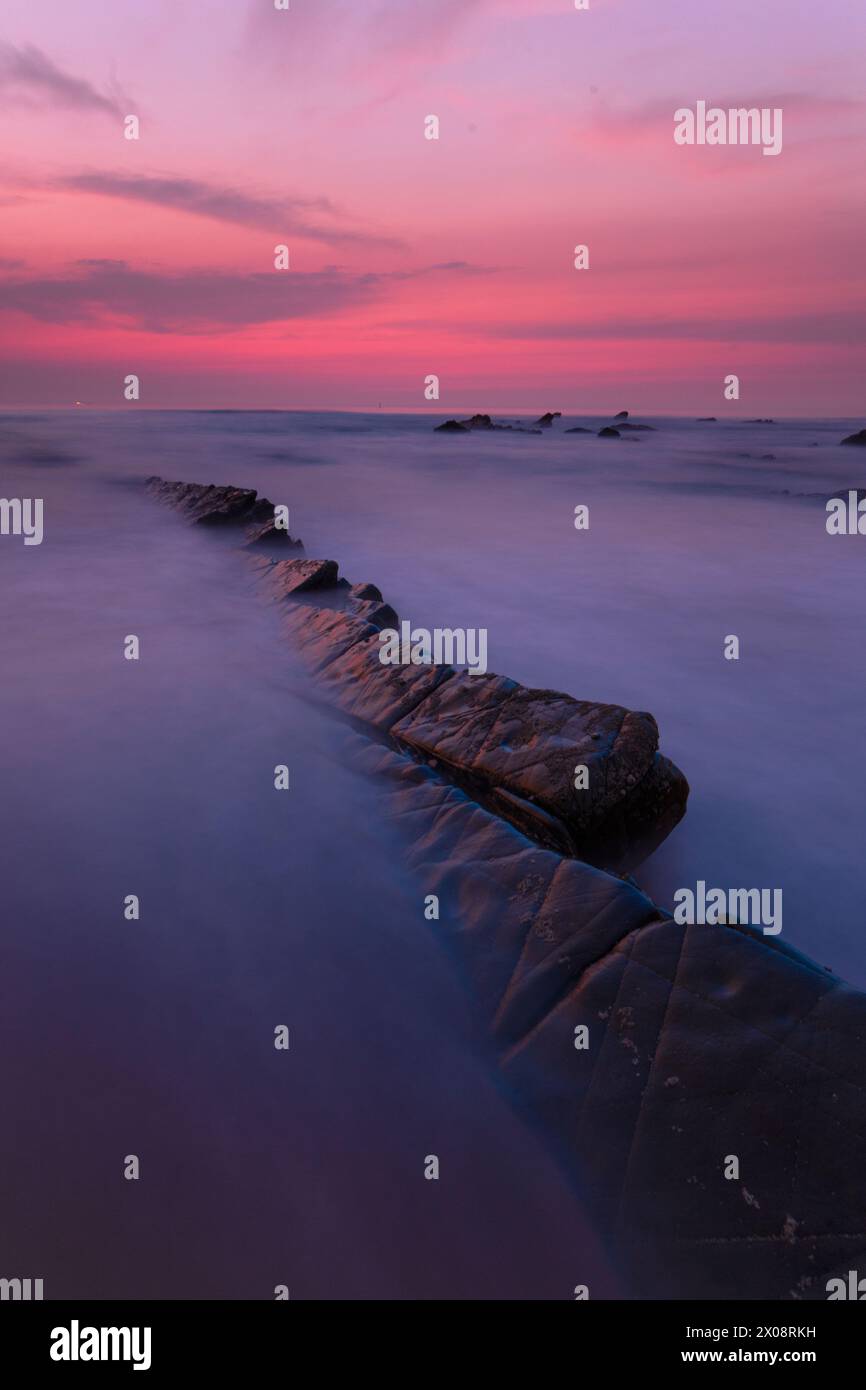 Un sentier rocheux naturel mène au cœur d'un coucher de soleil cramoisi à Playa de Barrika, Bilbao, avec les vagues de l'océan adoucies dans la brume Banque D'Images