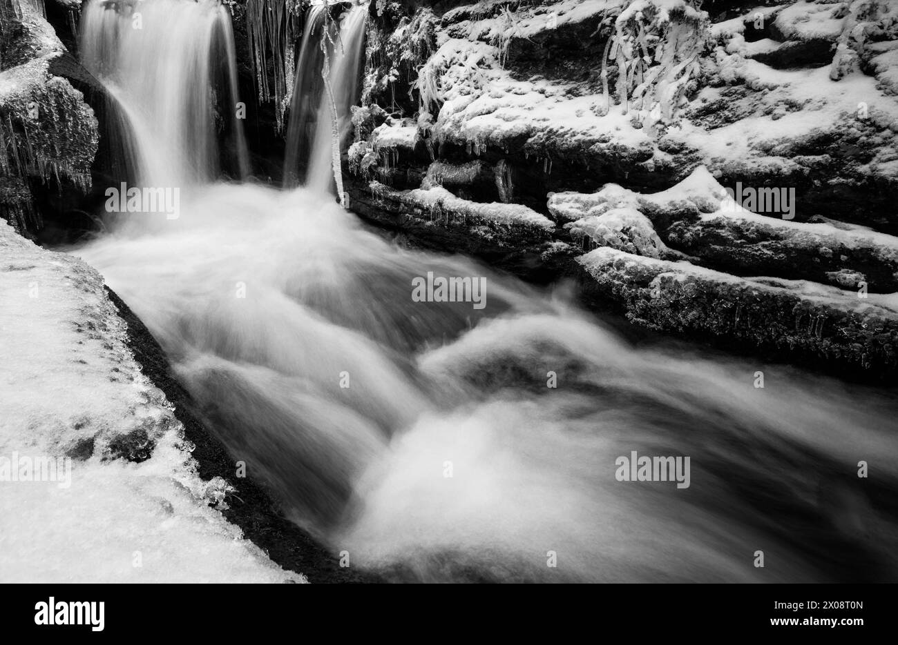 Une photo sereine en noir et blanc capturant une rivière qui coule à travers un paysage montagneux enneigé, avec une cascade en cascade et des rochers glacés. Banque D'Images