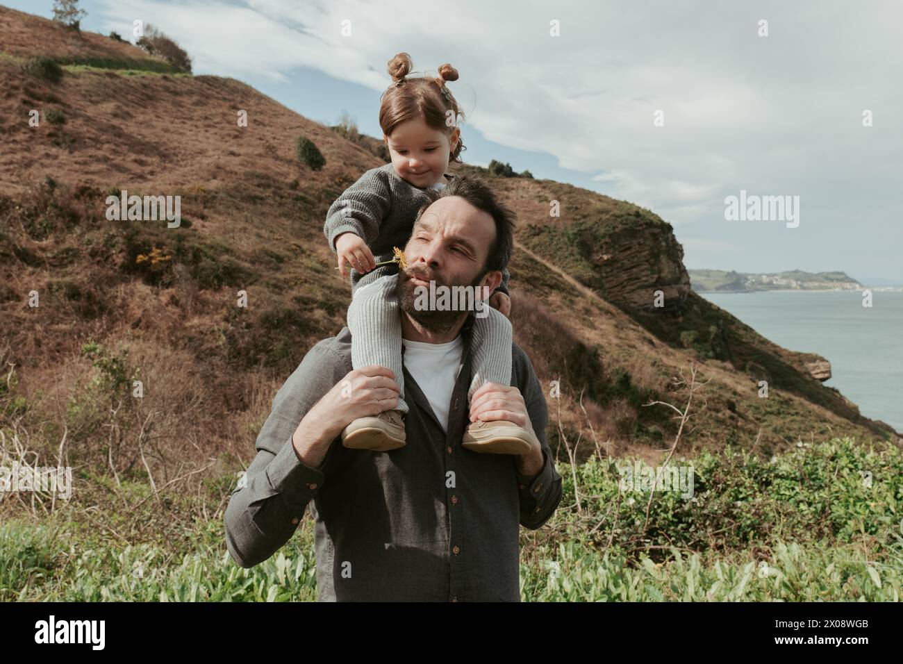 Un père aimant donne à sa jeune fille un tour d'épaule à l'extérieur, les deux profitant d'un paysage serein au bord de la mer Banque D'Images