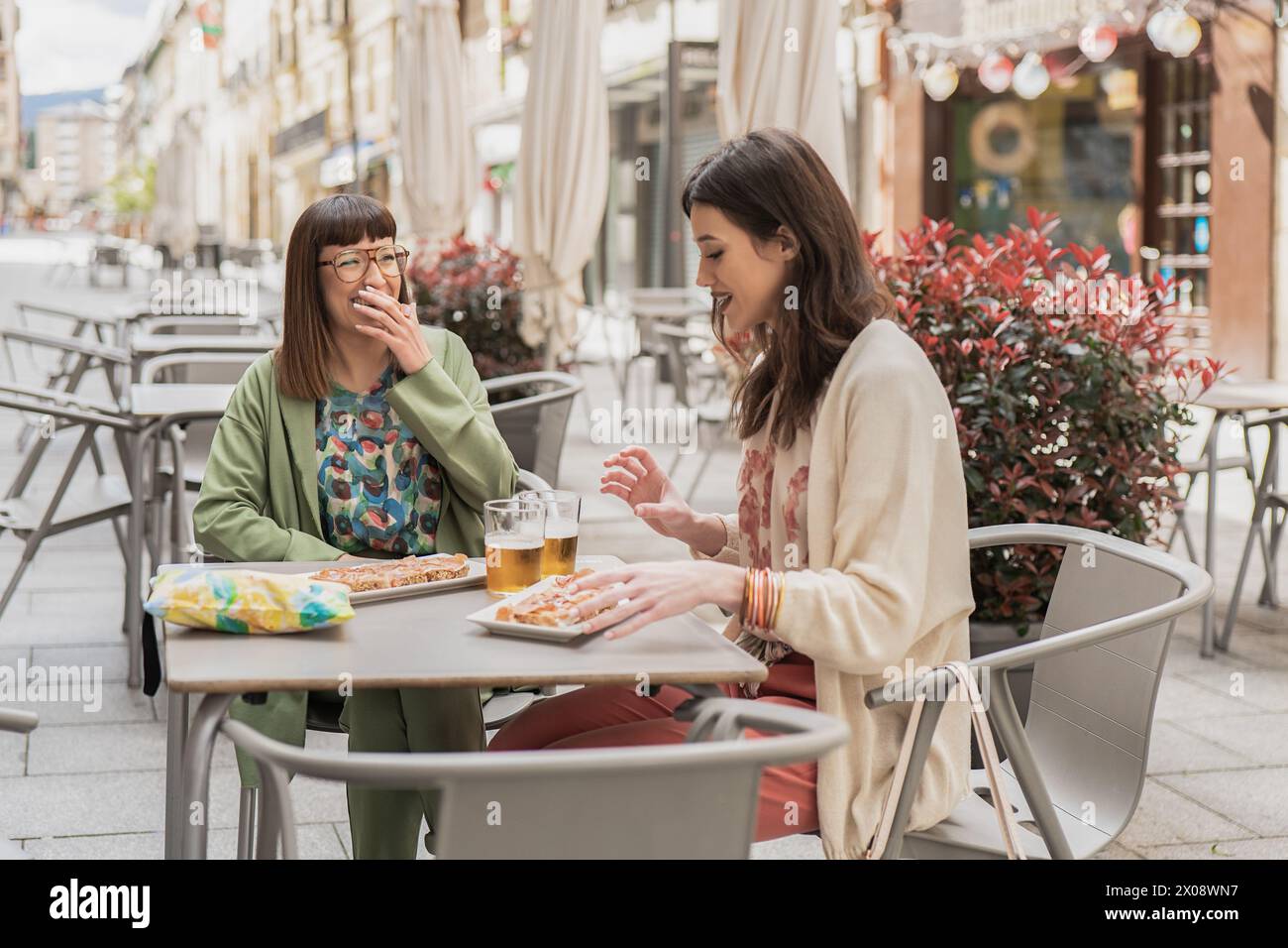 Une paire joyeuse de femmes partageant conversation et nourriture à une table de café sur le trottoir, incarnant l'essence d'une sortie printanière décontractée Banque D'Images