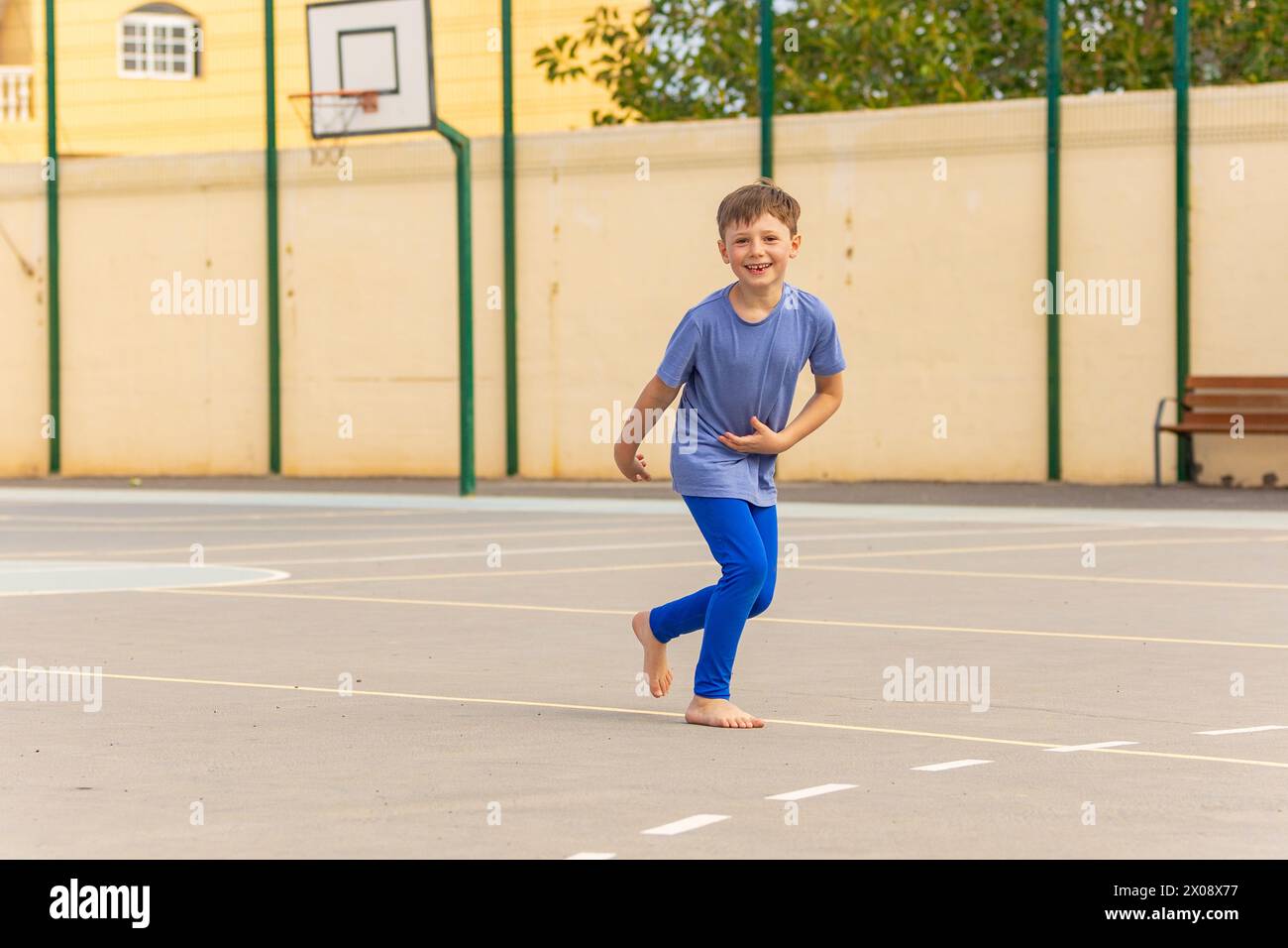 Un garçon heureux avec une expression joyeuse court pieds nus sur un terrain de basket-ball, portant une chemise bleue et un pantalon assorti. Banque D'Images