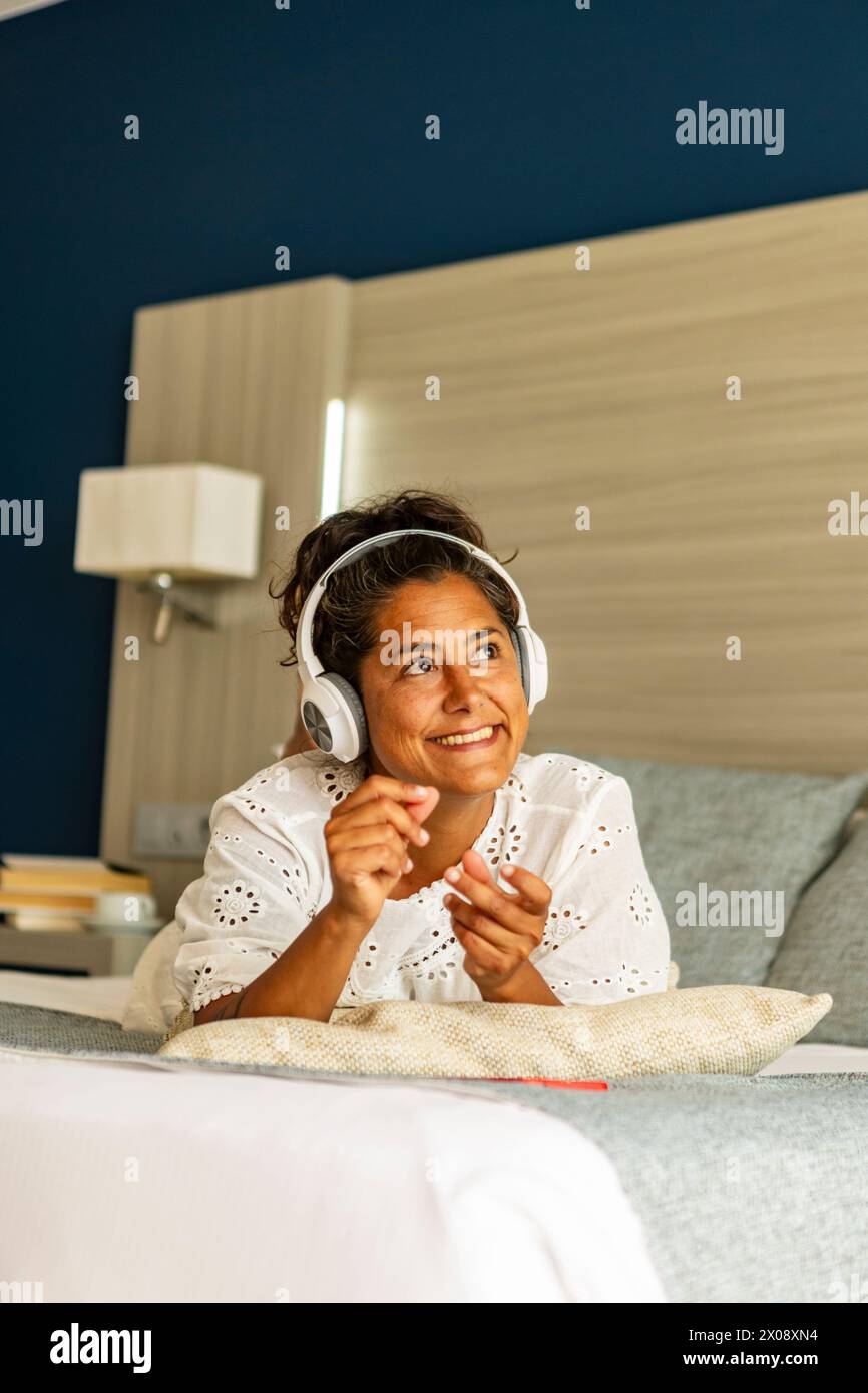 Une femme hispanique d'âge moyen souriante aime écouter de la musique avec des écouteurs, se prélasser confortablement sur un canapé à la maison, exsudant la détente décontractée et Banque D'Images