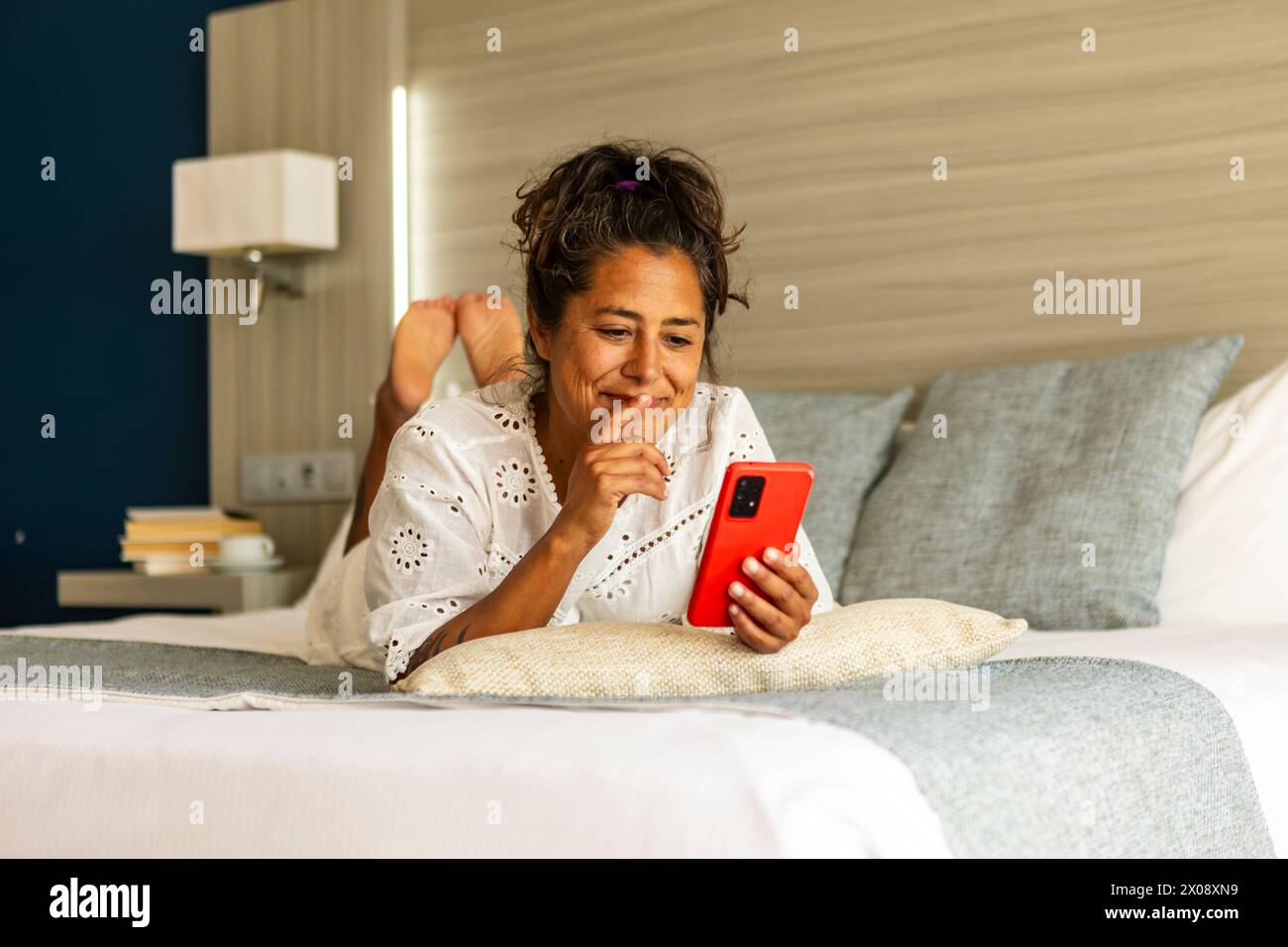 Une femme hispanique d'âge moyen souriante profite du temps libre tout en naviguant sur son smartphone, confortablement allongée sur un lit d'hôtel avec une décoration intérieure élégante Banque D'Images