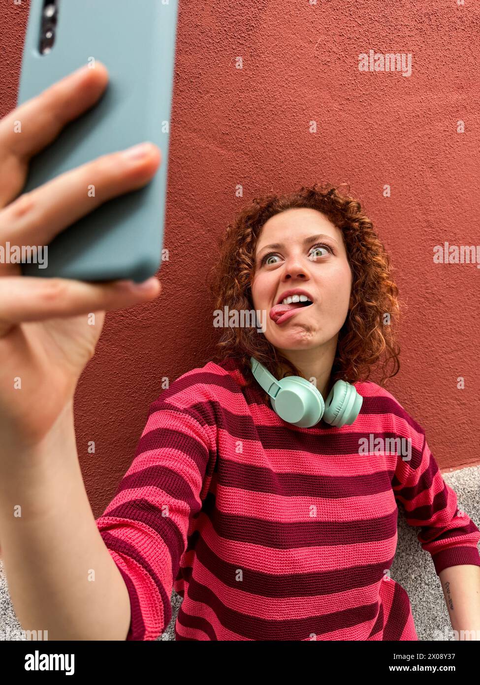 Une jeune femme rousse avec des cheveux bouclés et des écouteurs fait une grimace ludique alors qu'elle prend un selfie contre un mur rouge Banque D'Images