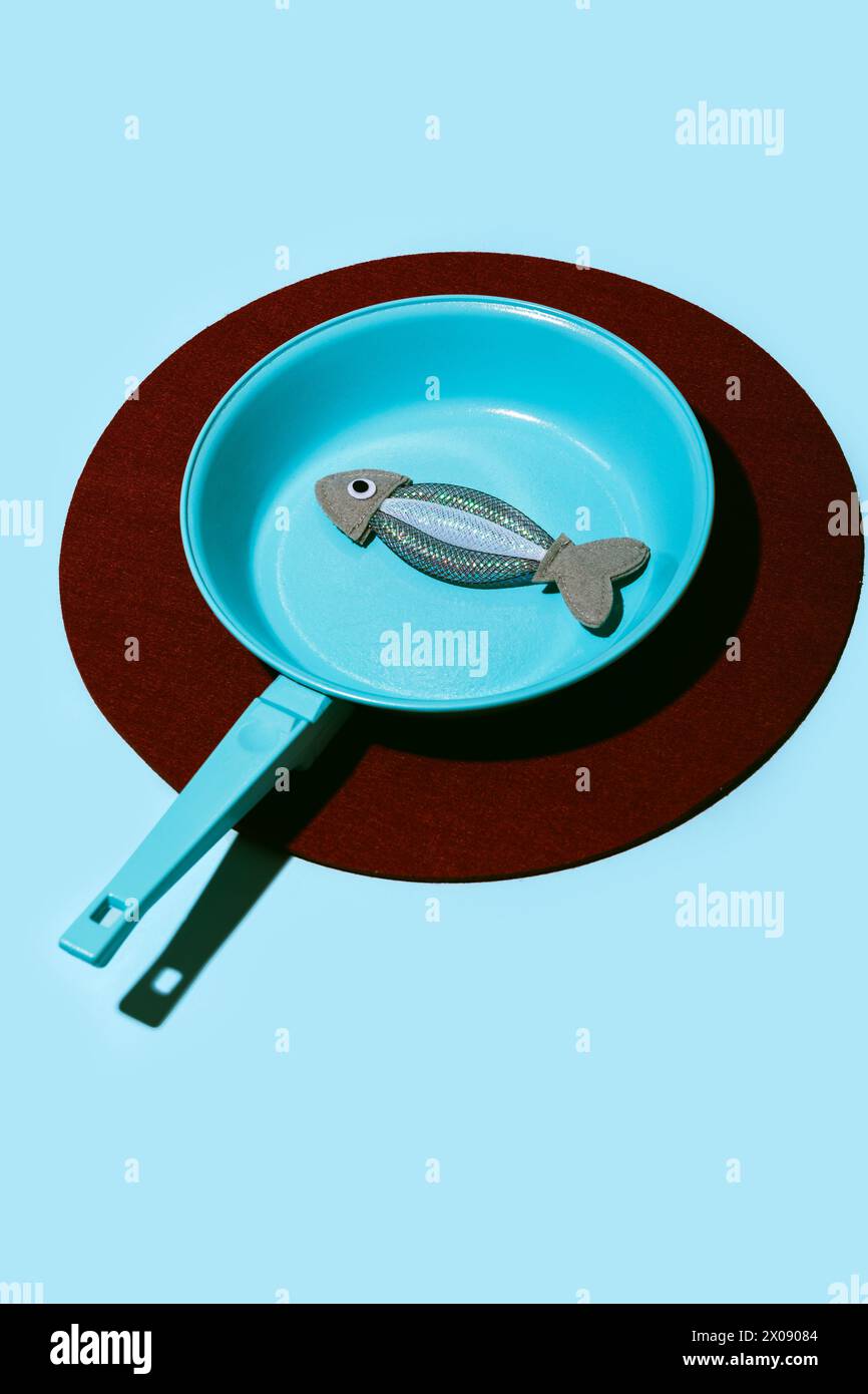 Une image conceptuelle représentant un poisson métallique sur une poêle sarcelle, projetant une ombre sur une surface bleue avec un contour circulaire brun. Banque D'Images