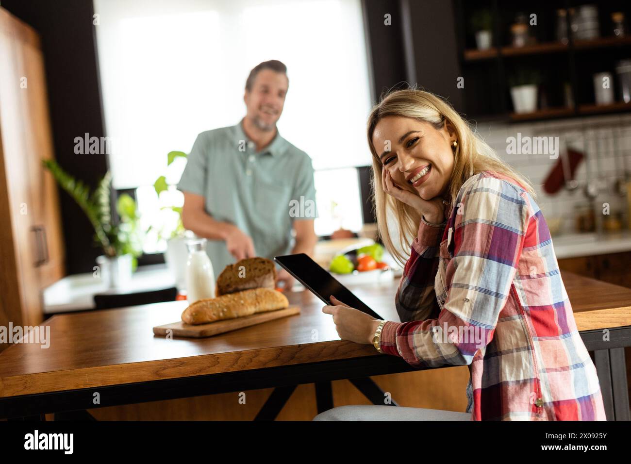 Une femme lit sur une tablette au comptoir de la cuisine tandis qu'un homme tenant une tomate lui sourit Banque D'Images