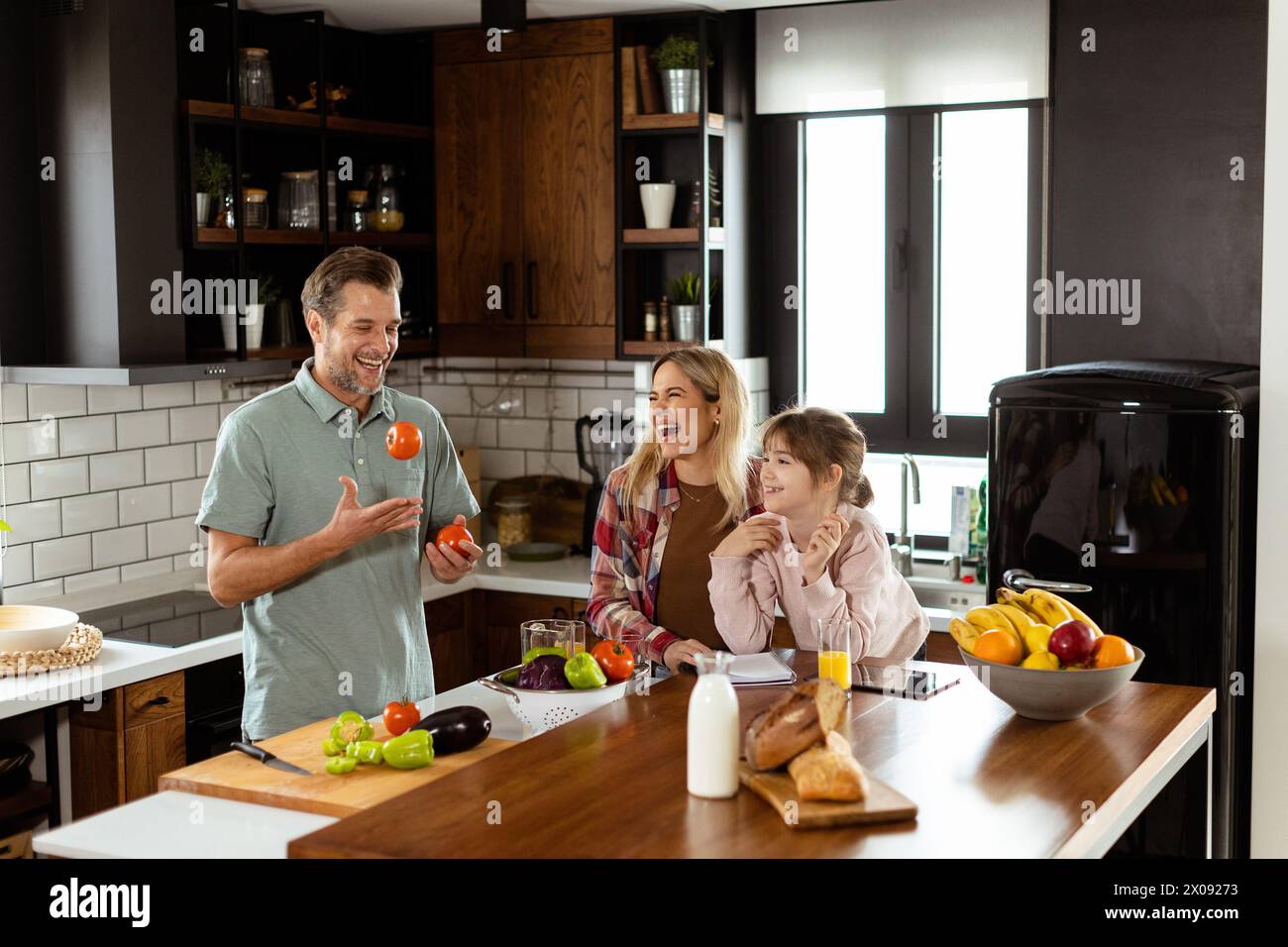 Jeune famille discutant et préparant la nourriture autour d'un comptoir de cuisine animé rempli d'ingrédients frais et d'ustensiles de cuisine Banque D'Images