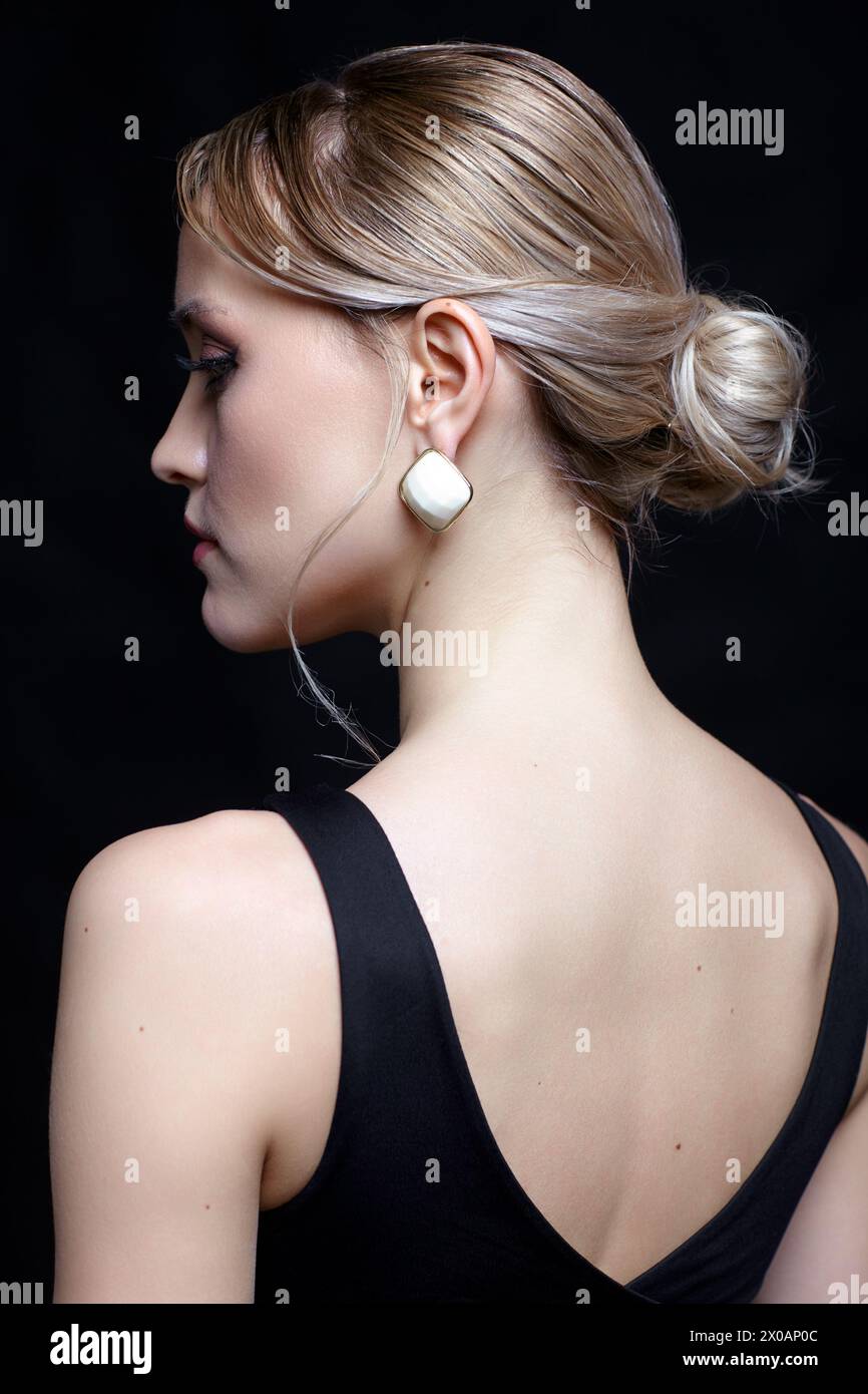 Portrait vue arrière de belle jeune femme blonde. Femme en t-shirt noir avec chignon coiffure sur fond noir. Banque D'Images