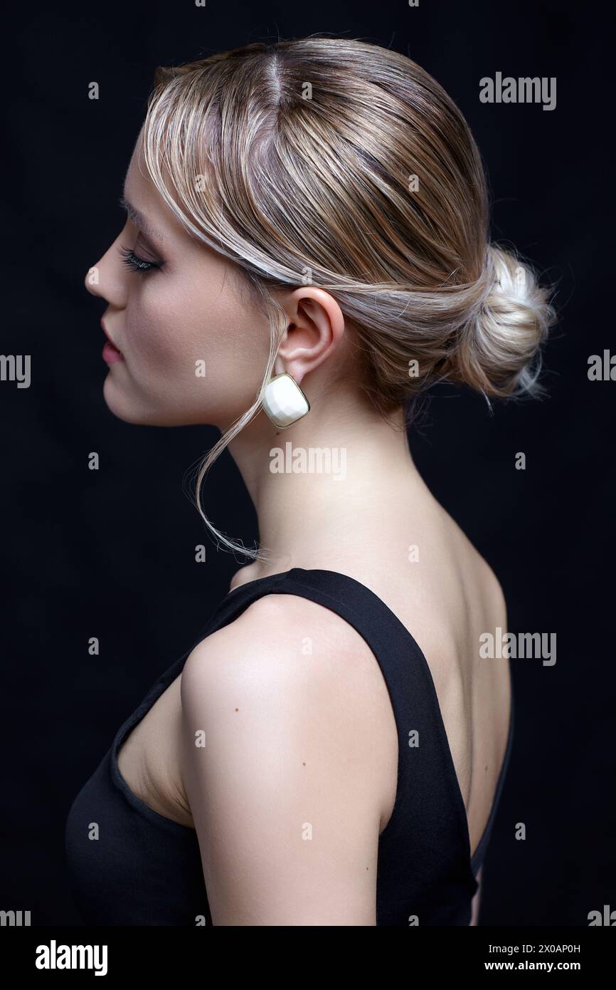 Portrait vue de côté de la belle jeune femme blonde. Femme en t-shirt noir avec chignon coiffure sur fond noir. Banque D'Images