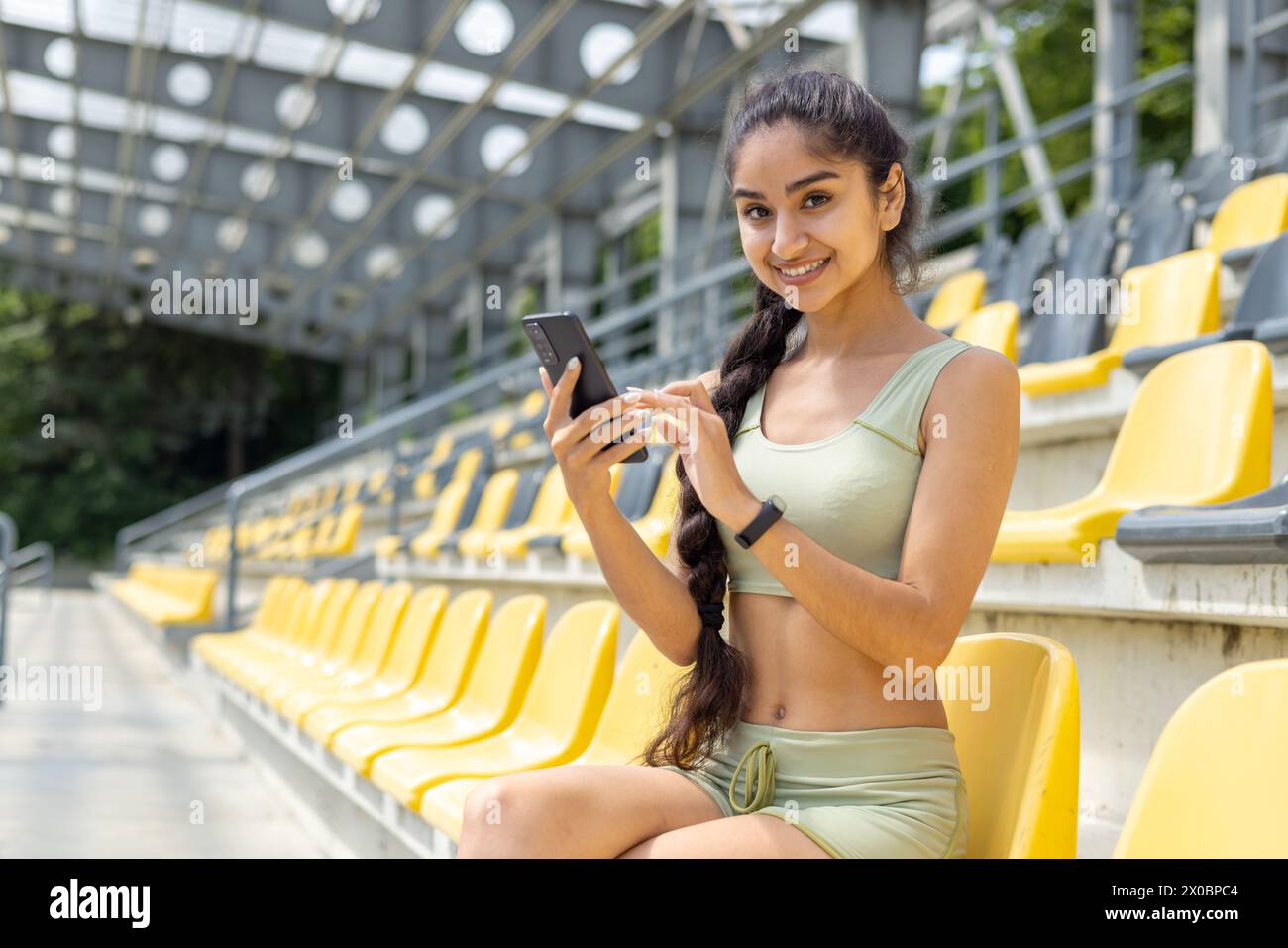 Portrait d'une jeune femme indienne assise en vêtements de sport sur la tribune du stade, tenant un téléphone portable et souriant à la caméra. Banque D'Images