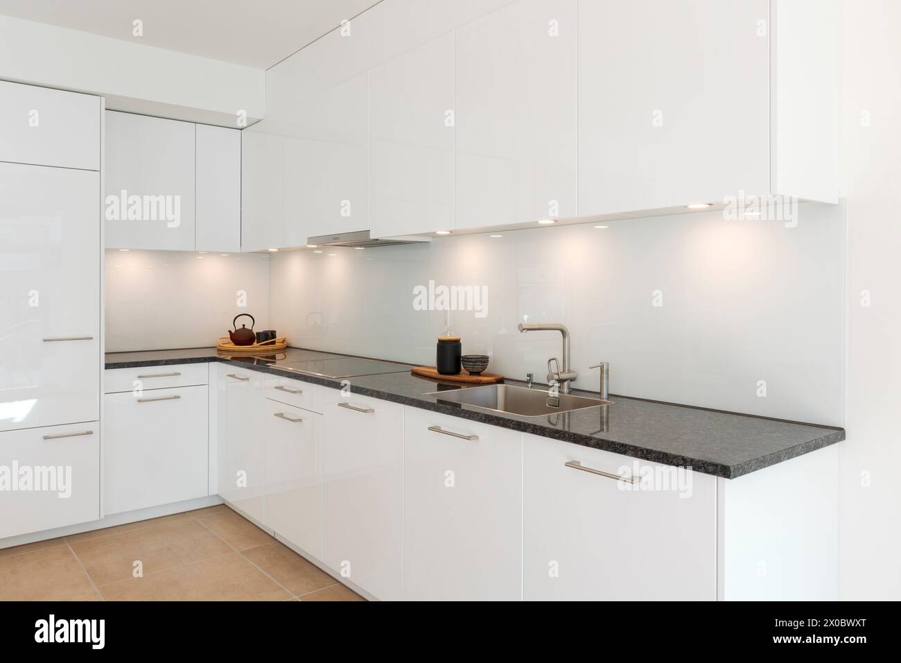 Cuisine moderne minimaliste blanc brillant avec un comptoir en marbre noir. Des spots éclairent le comptoir, au-dessus duquel se trouvent deux plateaux, dont un avec un thé Banque D'Images