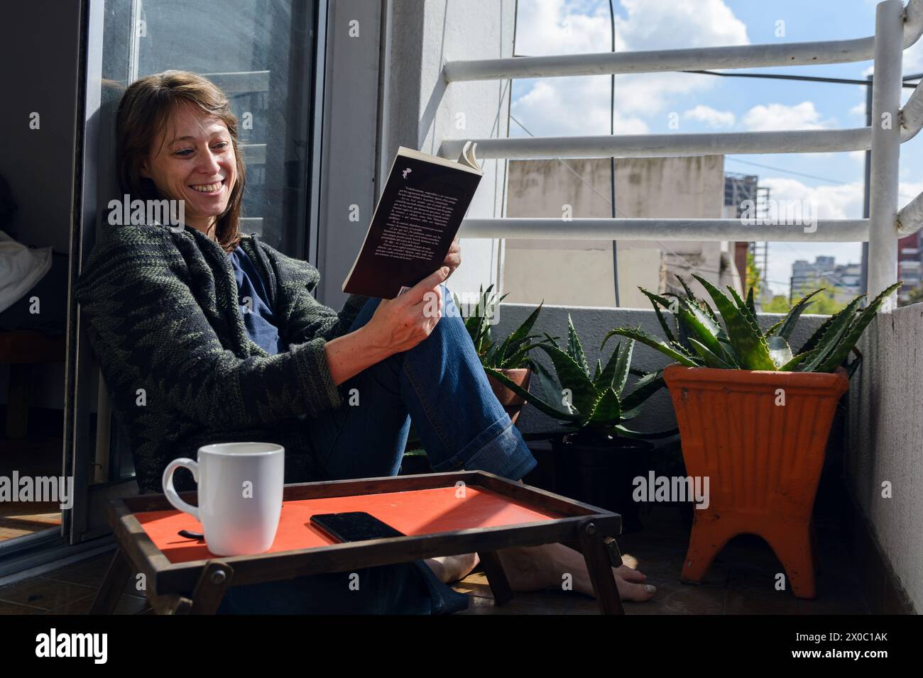 femme adulte latine heureuse avec des vêtements froids, à la maison assis sur la terrasse avec des plantes de savila, livre de lecture très calmement et boire du café, lire le roman, Banque D'Images
