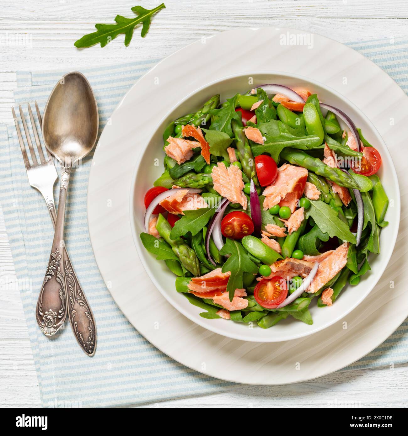 salade de saumon fumé avec asperges, salade de roquette, petits pois, tomates et oignon rouge dans un bol blanc sur une table en bois blanc avec fourchette et cuillère, horizontont Banque D'Images