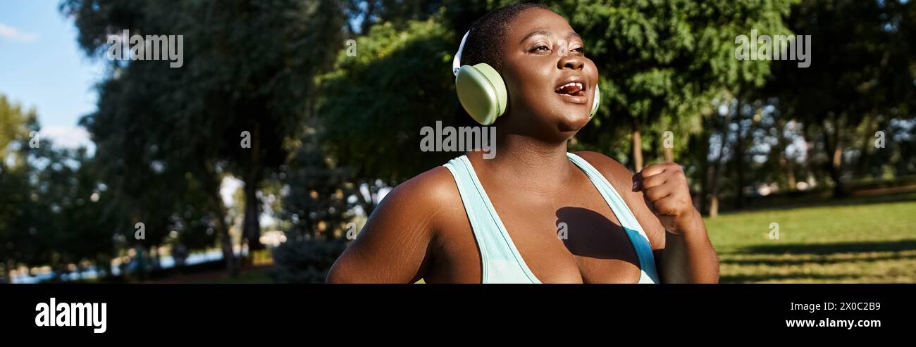 Une femme afro-américaine en vêtements de sport, avec une attitude positive du corps, debout dans un parc tout en portant des écouteurs. Banque D'Images