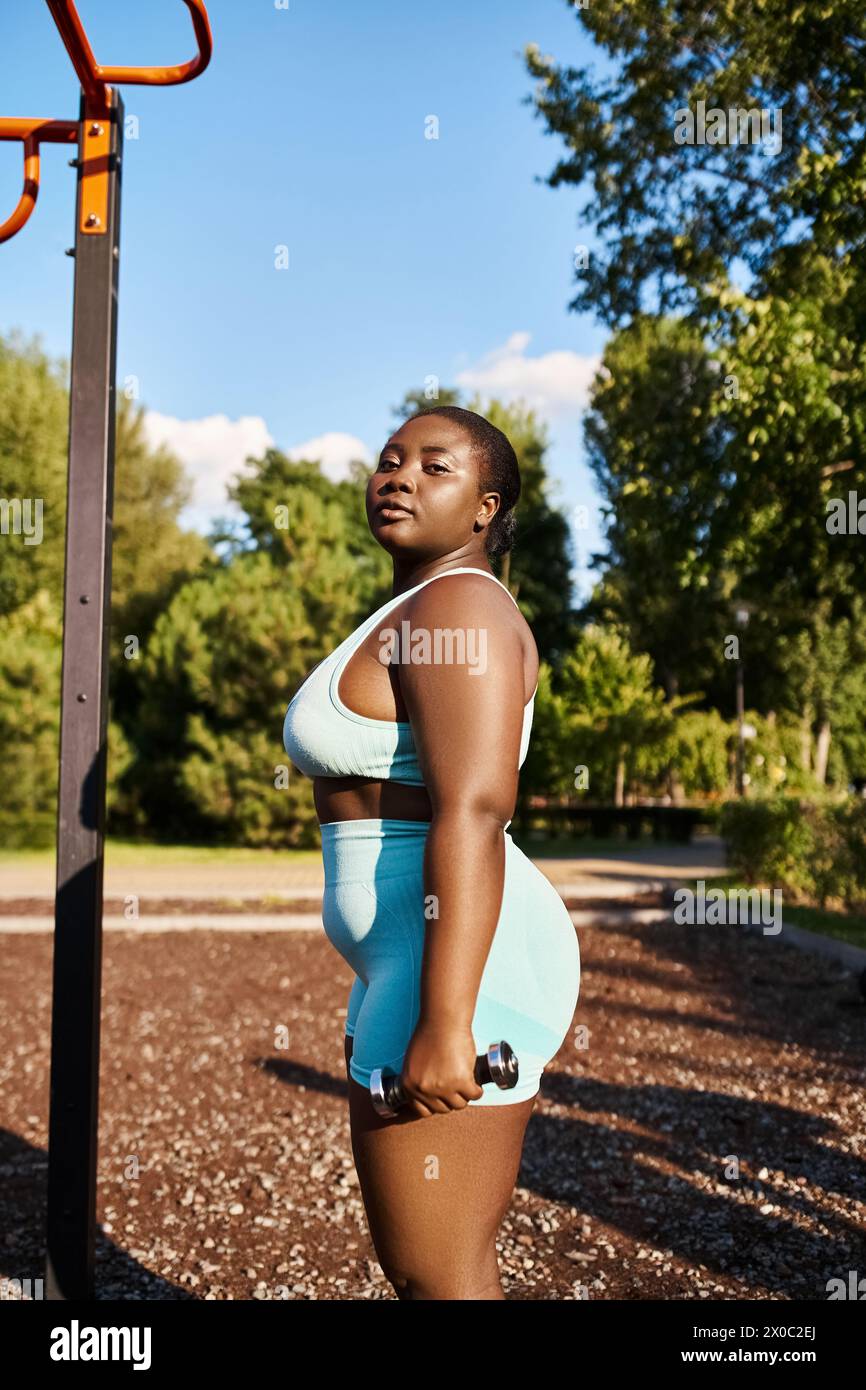 Une femme afro-américaine sinueuse en vêtements de sport se tient à côté d'un poteau dans un parc. Banque D'Images