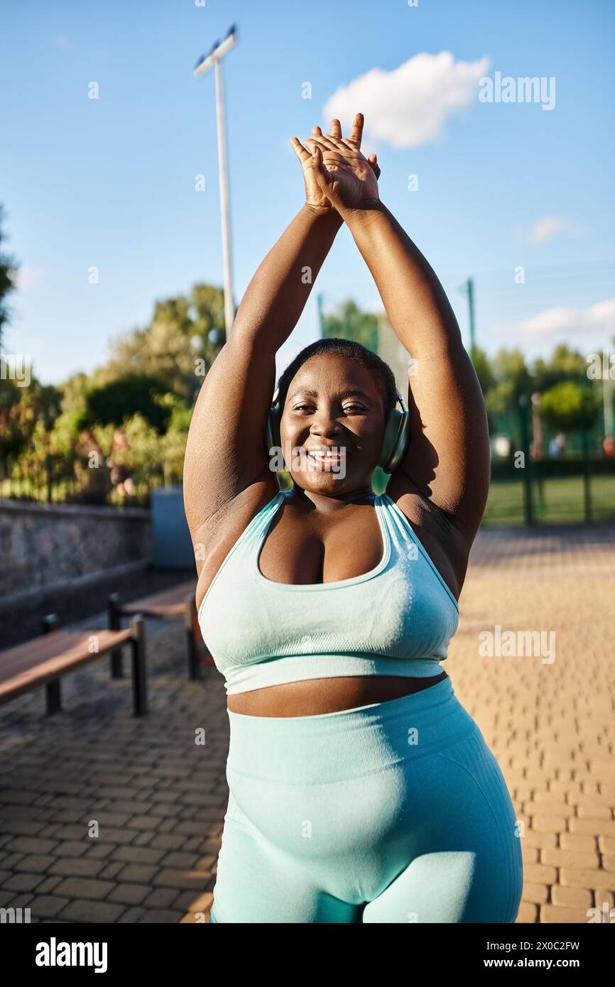 Une femme afro-américaine avec une attitude positive du corps, portant un haut de soutien-gorge de sport bleu, étire ses bras gracieusement à l'extérieur. Banque D'Images