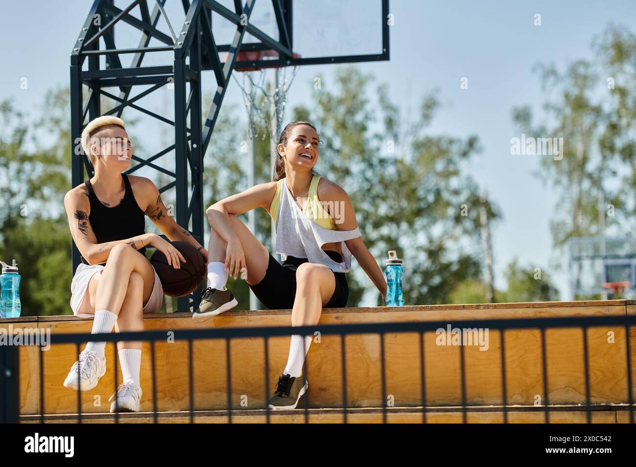 Deux jeunes femmes, amies athlétiques, assises sur un banc devant un panier de basket-ball par une journée ensoleillée. Banque D'Images
