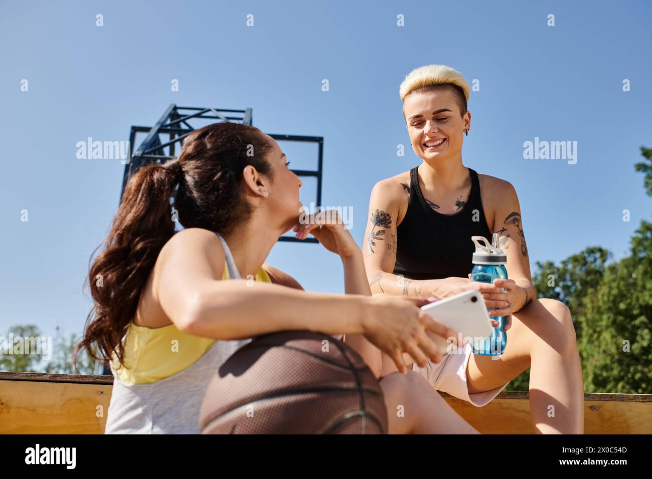 Deux jeunes femmes athlétiques sont assises sur le sol, engagées dans la conversation, profitant d'un moment de connexion alors qu'elles prennent une pause de jouer au basket-ball. Banque D'Images