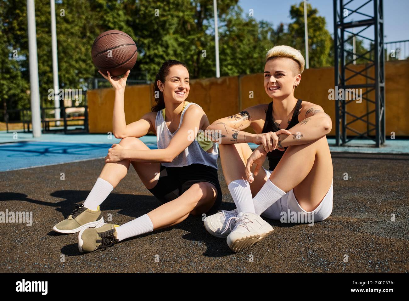 Deux jeunes femmes, athlétiques et animées, assises sur le sol avec un ballon de basket entre elles, profitant d'une journée d'été ensoleillée. Banque D'Images