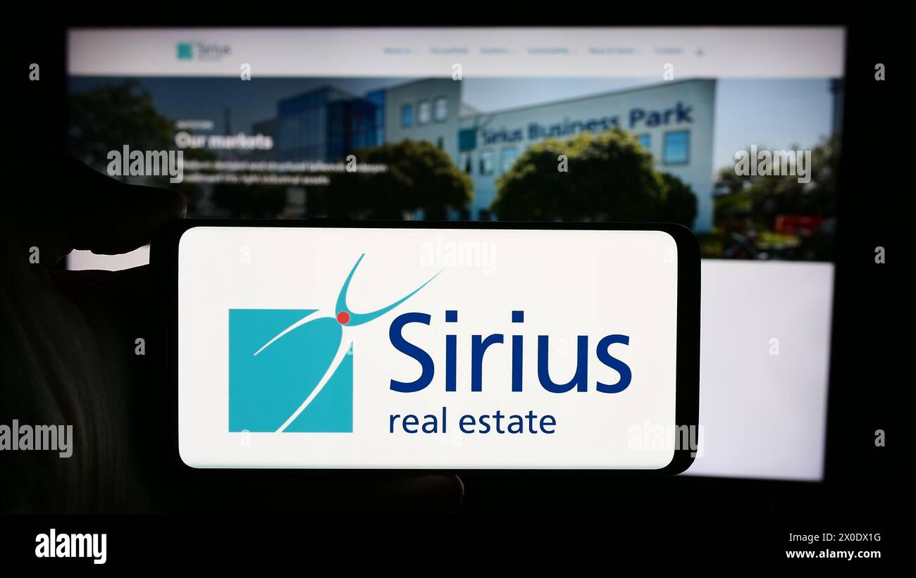 Personne tenant le téléphone portable avec le logo de la société de propriété commerciale Sirius Real Estate Limited en face de la page Web. Concentrez-vous sur l'affichage du téléphone. Banque D'Images