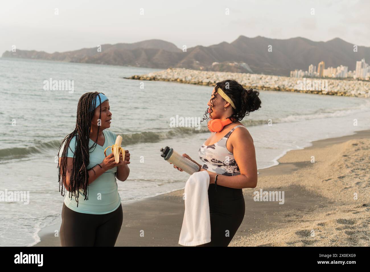 Deux femmes en équipement d'entraînement prennent une pause collation sur la plage, l'une mangeant une banane et l'autre tenant une bouteille d'eau. Banque D'Images