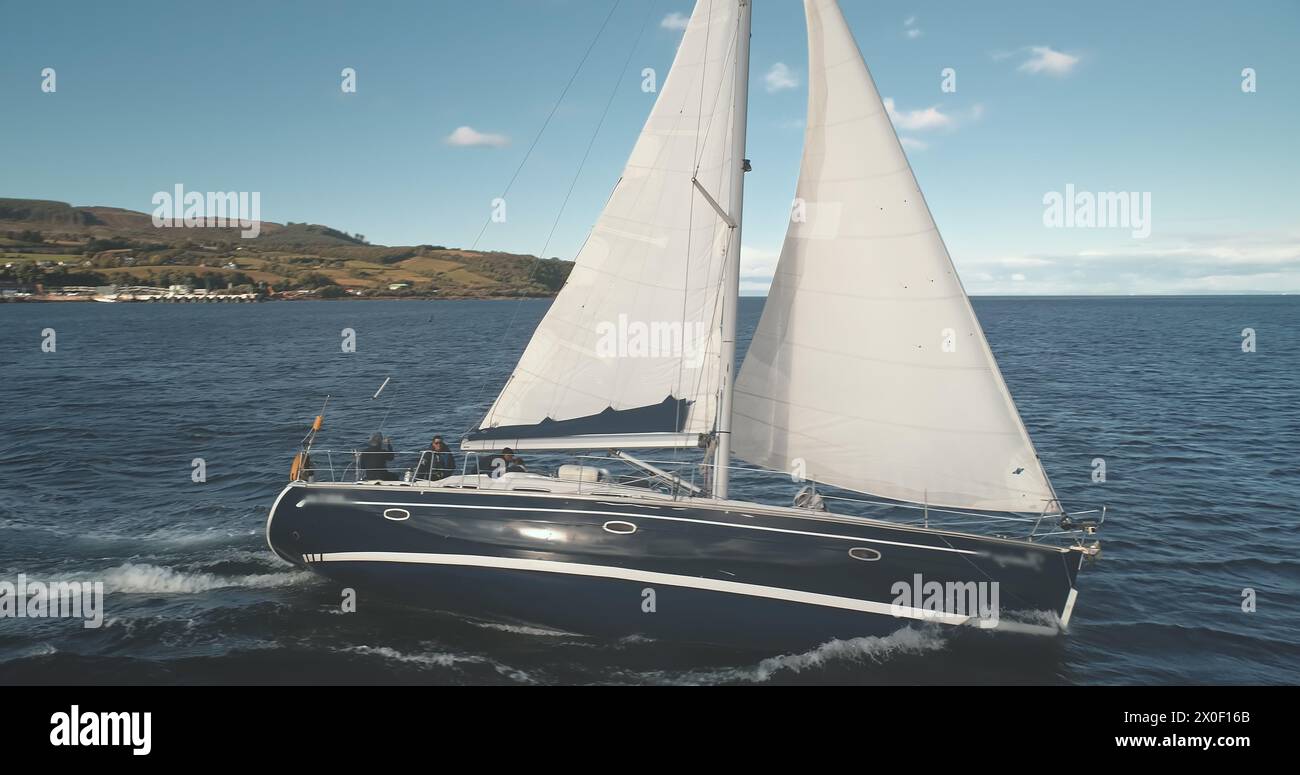 Reflet du soleil au yacht de luxe en gros plan sur la baie aérienne de l'océan. Voiles blanches au vent sur le bateau à passagers. Course de régates en pleine mer. Paysage marin serein et calme du golfe de Brodick, île d'Arran, Écosse, Europe Banque D'Images