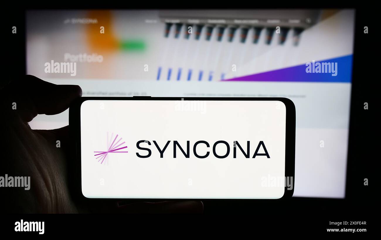 Personne tenant le téléphone portable avec le logo de la société d'investissement britannique Syncona Limited en face de la page Web de l'entreprise. Concentrez-vous sur l'affichage du téléphone. Banque D'Images