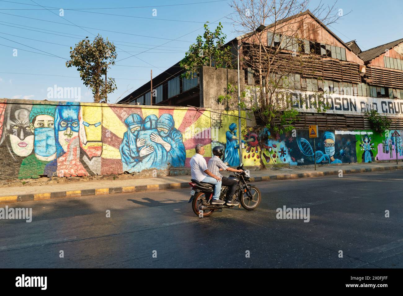 Un motocycliste avec passager passe un mur à Byculla, Mumbai, Inde, peint avec un certain nombre de peintures murales colorées avec des thèmes COVID-19 Banque D'Images