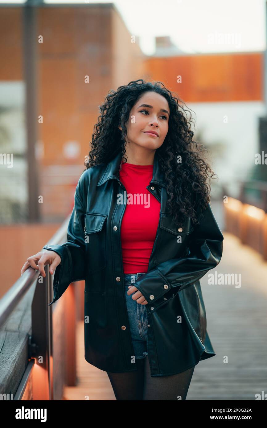Femme élégante aux cheveux bouclés en tenue décontractée s'appuyant sur une balustrade au crépuscule Banque D'Images