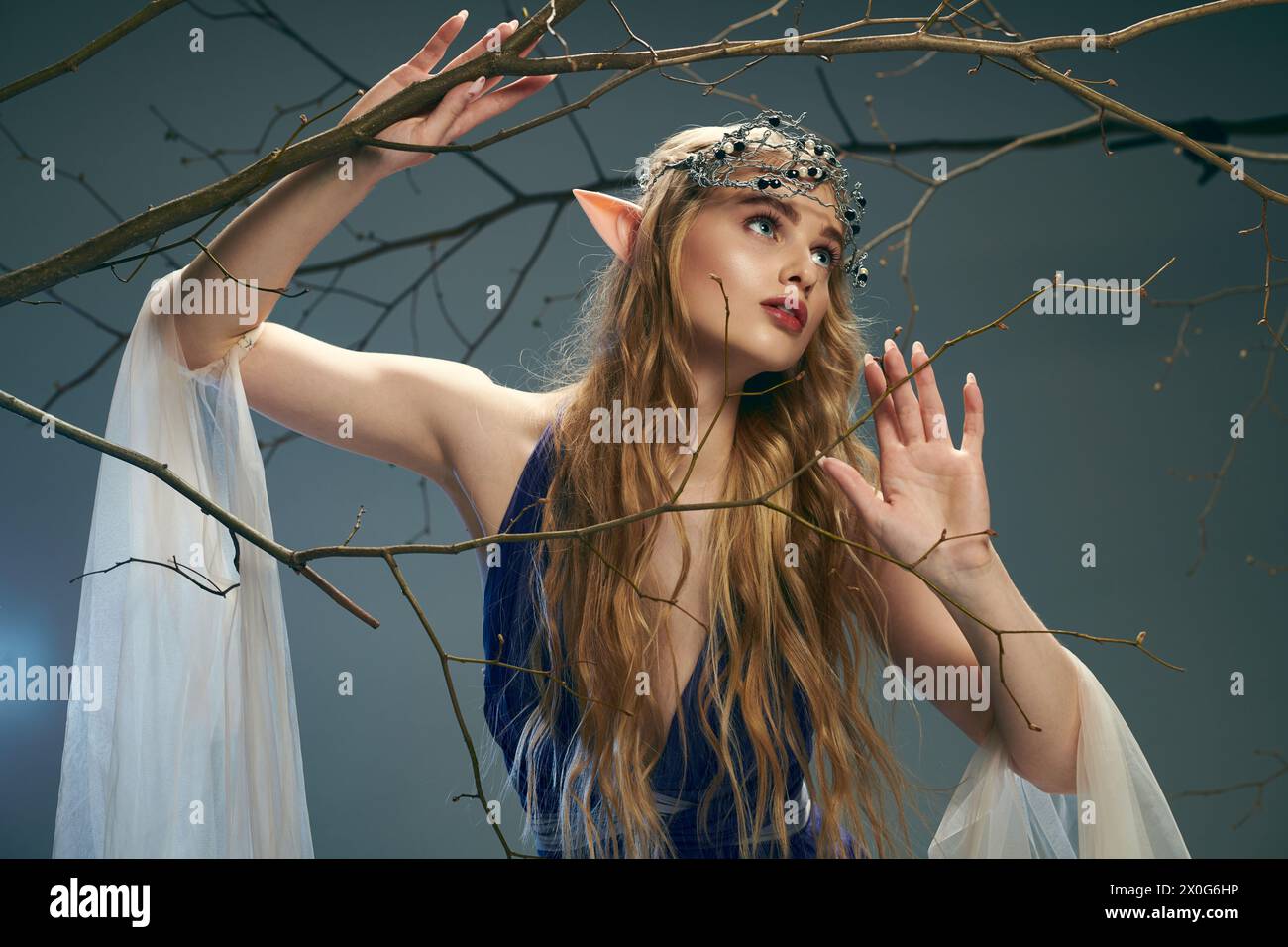 Une jeune femme portant une robe bleue fluide se tient gracieusement à côté d'un arbre dans un cadre fantastique magique. Banque D'Images