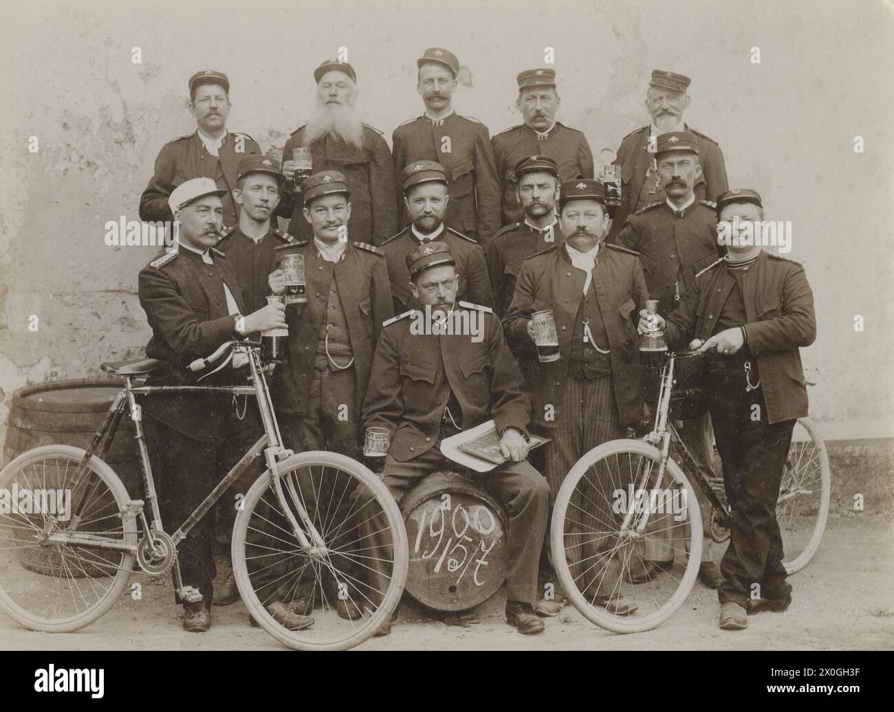 Un groupe de 14 serviteurs dans leur posture uniforme pour une photo. Ils portent des casquettes et tiennent un mug à bière dans leurs mains. Le secrétaire est assis pour une bière. Les deux hommes sur le côté tiennent leurs vélos. Un des hommes est fumeur, beaucoup portent des montres de poche. La date du jour de la photo est inscrite sur le mug à bière. [traduction automatique] Banque D'Images