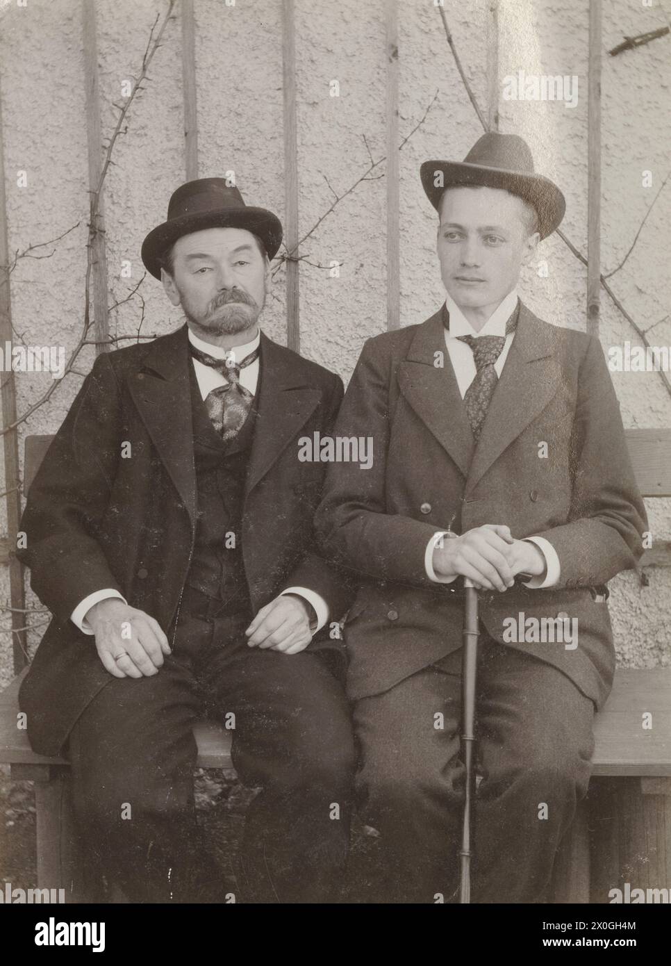 2 hommes en costumes assis sur un banc en bois devant un mur de maison blanc avec un treillis en bois (encore sans feuilles). Le plus âgé, âgé de 40-45 ans, avec une barbe courte et taillée, a un col blanc debout et ses mains reposent sur ses cuisses, le plus jeune homme plus grand, âgé d'environ 20 ans, a ses mains reposant sur un bâton de marche entre ses jambes. Il regarde tristement devant la caméra, à l'automne ou au printemps [traduction automatique] Banque D'Images