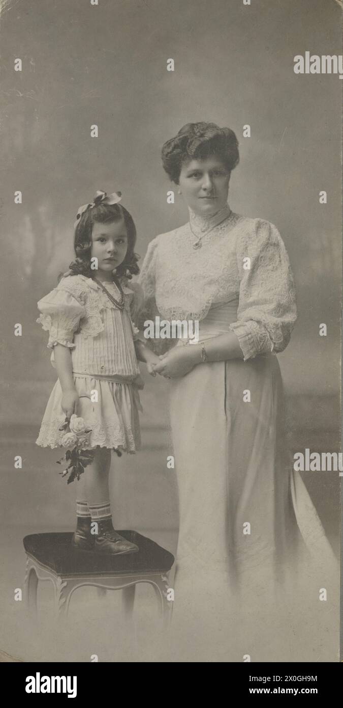 Photo de Johanna König avec sa fille Elsa, une femme de la classe moyenne d'environ 30 ans, vêtue d'une robe blanche et d'un haut en dentelle, d'une chaîne et d'un bracelet en or. À côté d'elle se trouve sa fille debout sur un tabouret recouvert de tissu avec une robe blanche en dentelle longueur genou, un collier, un noeud de cheveux et des roses blanches dans sa main [traduction automatique] Banque D'Images