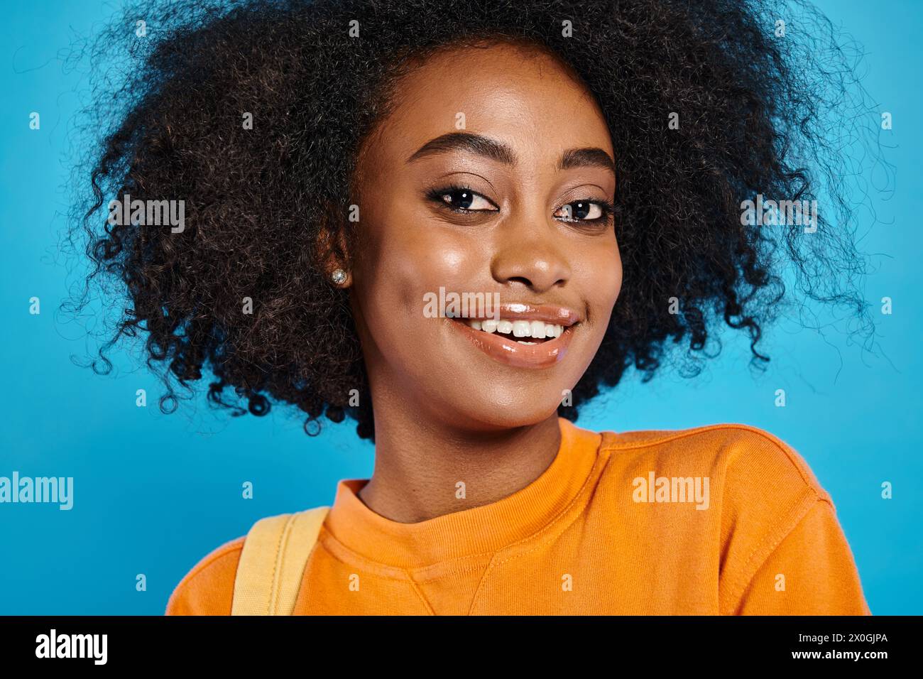 Une fille d'université afro-américaine avec une coiffure afro magnifique sourit brillamment dans une tenue décontractée sur un fond de studio bleu. Banque D'Images