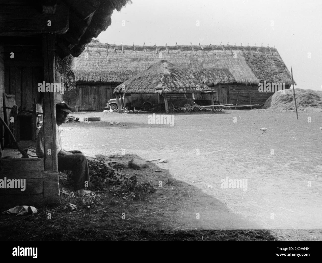 Un agriculteur est assis à l'ombre d'une ferme. Les poulets courent autour de la cour, les chariots sont garés sous la grange. Un camion de la Wehrmacht est garé devant le hangar de chaume. La photo a été prise par un membre du Radfahrgrenadierregiment 2 / Radfahrsicherungsregiment 2, sur le front de l'est. [traduction automatique] Banque D'Images